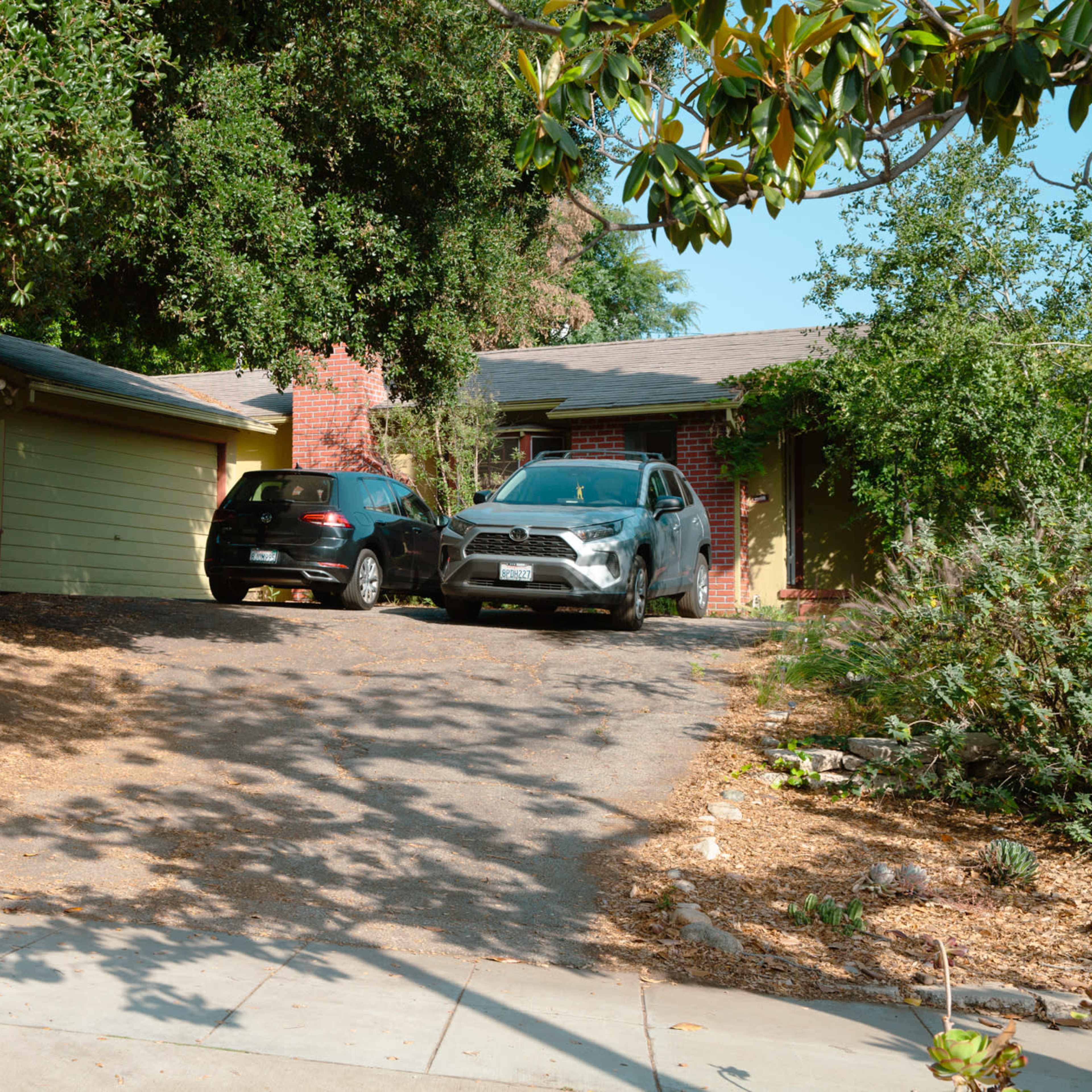 A driveway leads to a house with a green exterior, flanked by two parked cars and surrounded by trees and gravel.