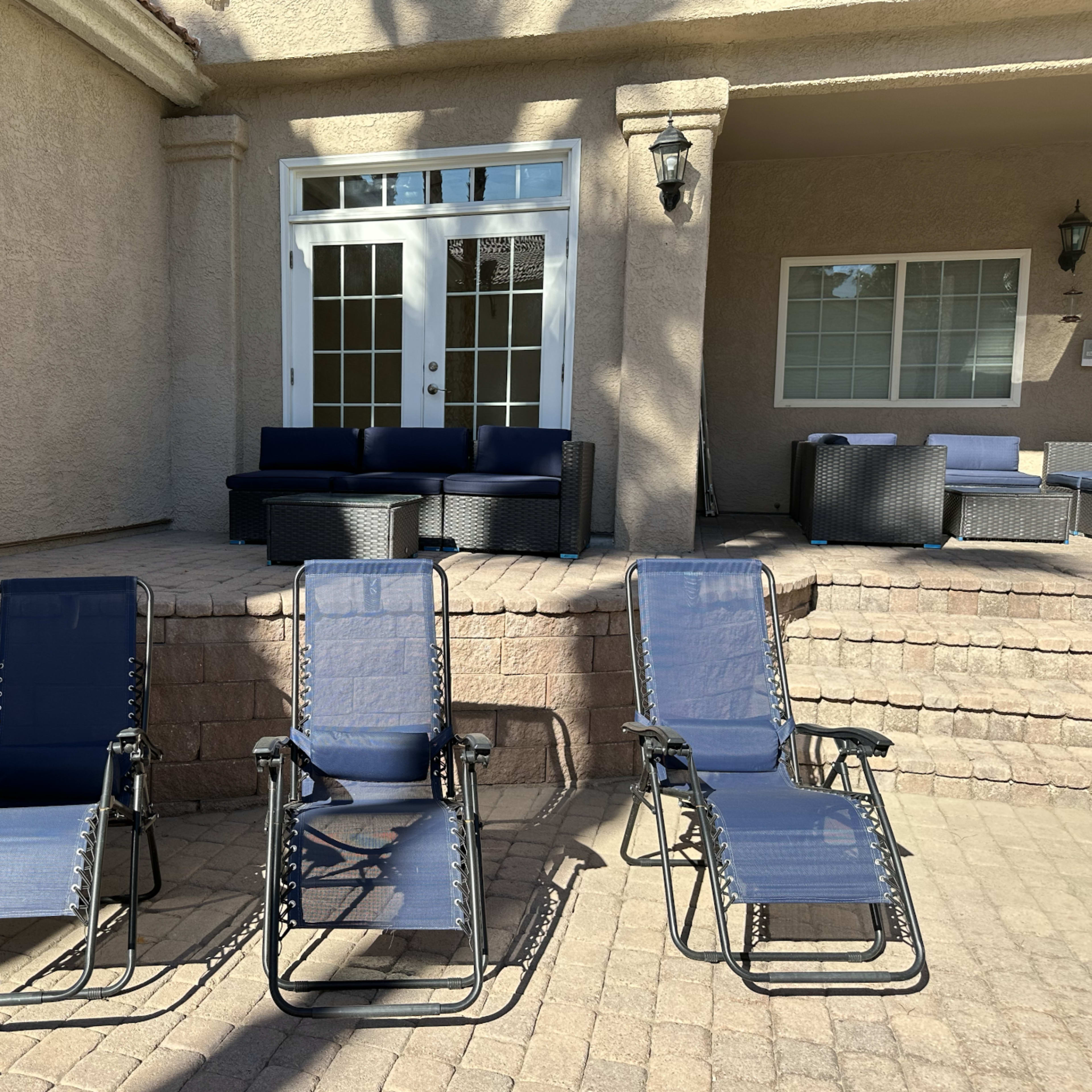 Three blue lounge chairs are positioned in front of a light-colored stone terrace leading to a building with large glass doors and a seating area.