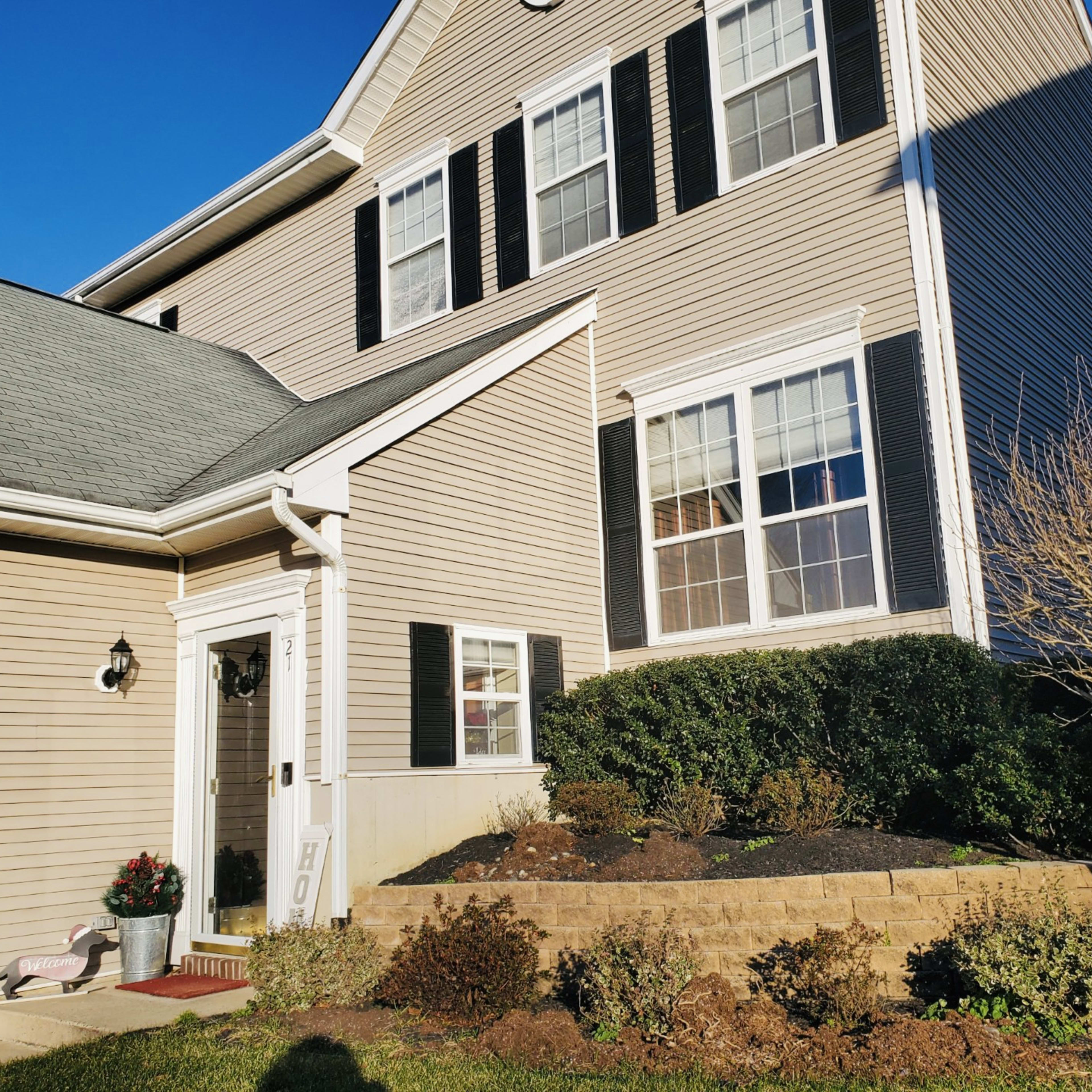 A two-story house with beige siding and black shutters features a front porch and landscaped garden.
