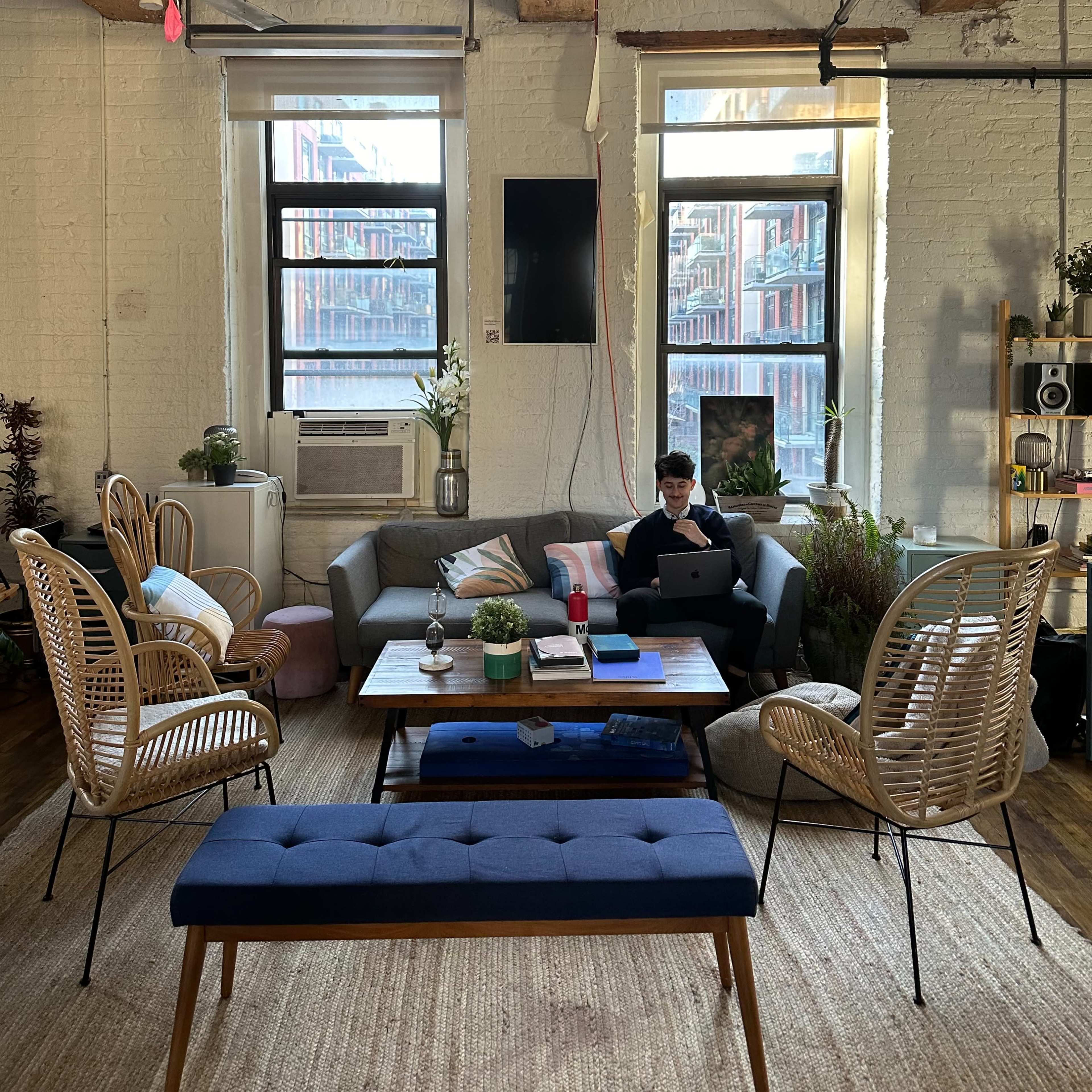 A person sits on a couch with a laptop in a spacious living area featuring large windows, plants, and wooden beams in the ceiling.