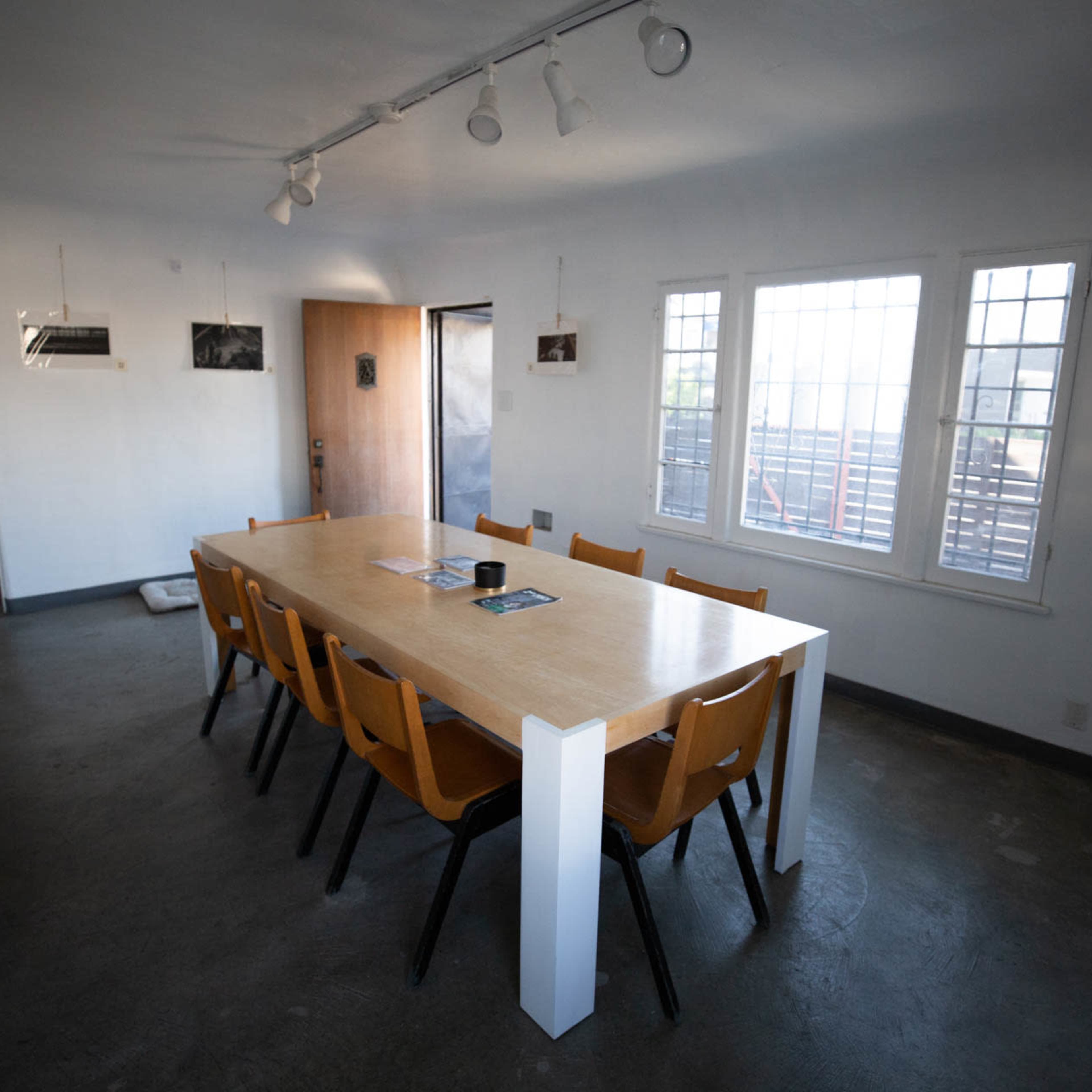 A long wooden table with eight chairs is positioned in a well-lit room featuring large windows and minimal decorations.