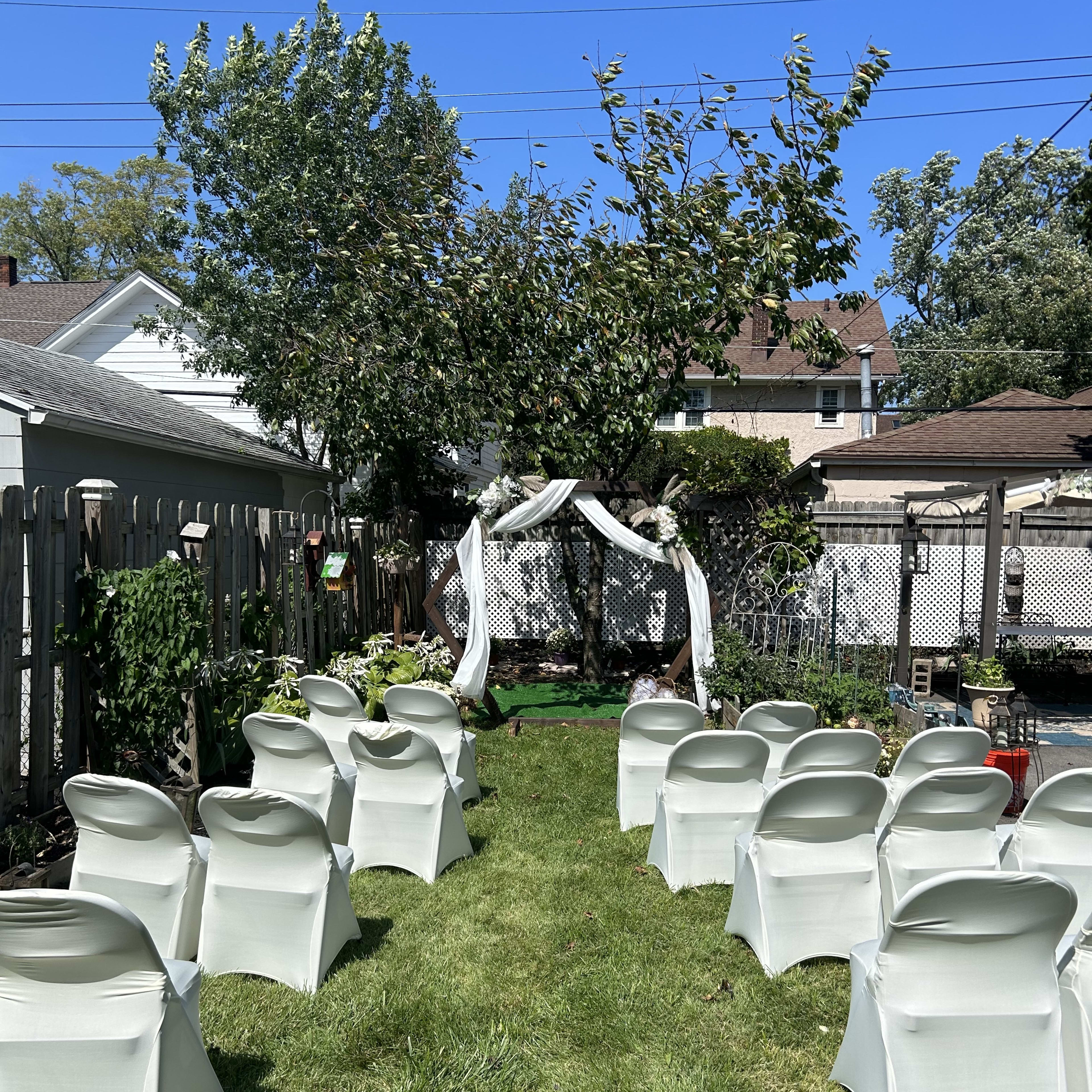 A backyard is set up with white chairs facing a decorative arch under a tree for an outdoor event.