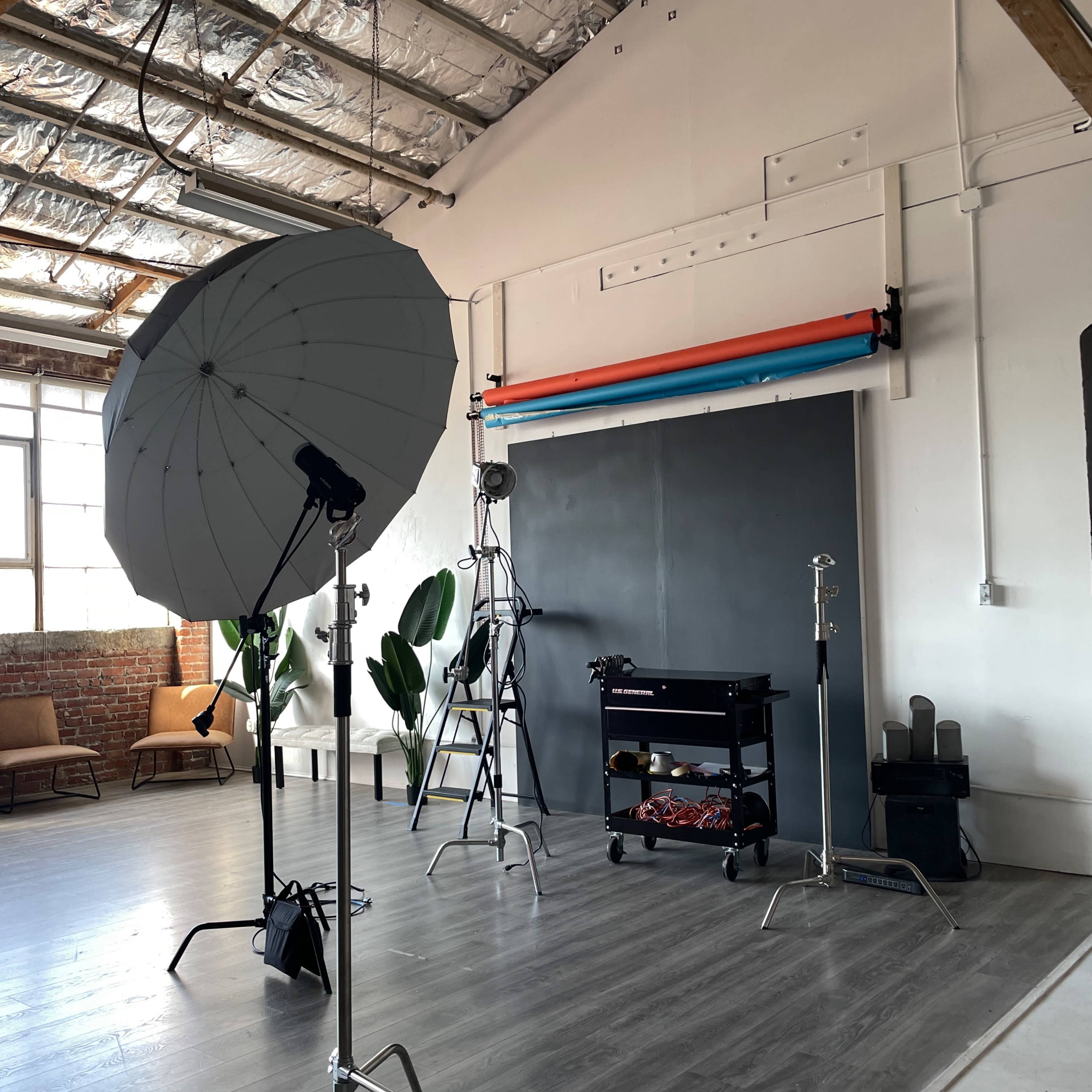 A photography studio equipped with softbox lighting, a backdrop stand, and various props, set against a backdrop of large windows and exposed brick walls.