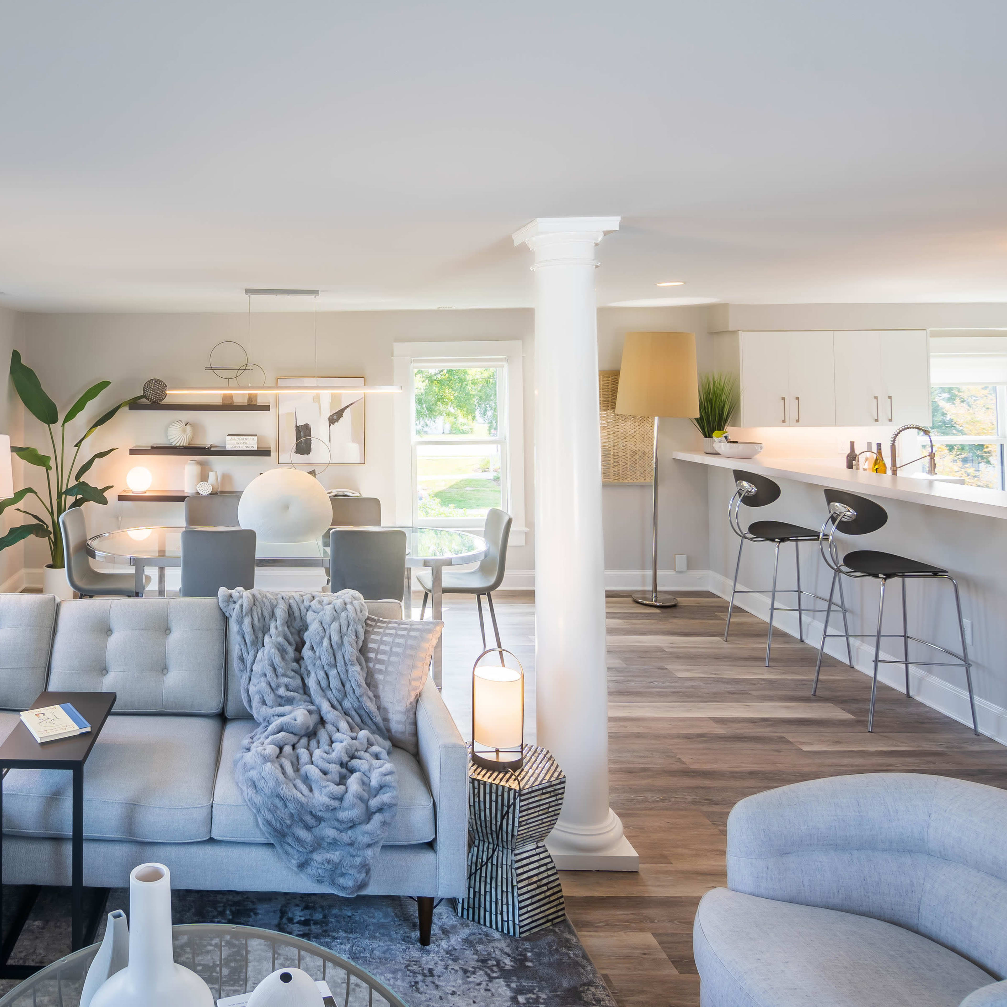A modern living room features a light-colored sectional sofa, a coffee table, and a dining area with bar stools in the background.
