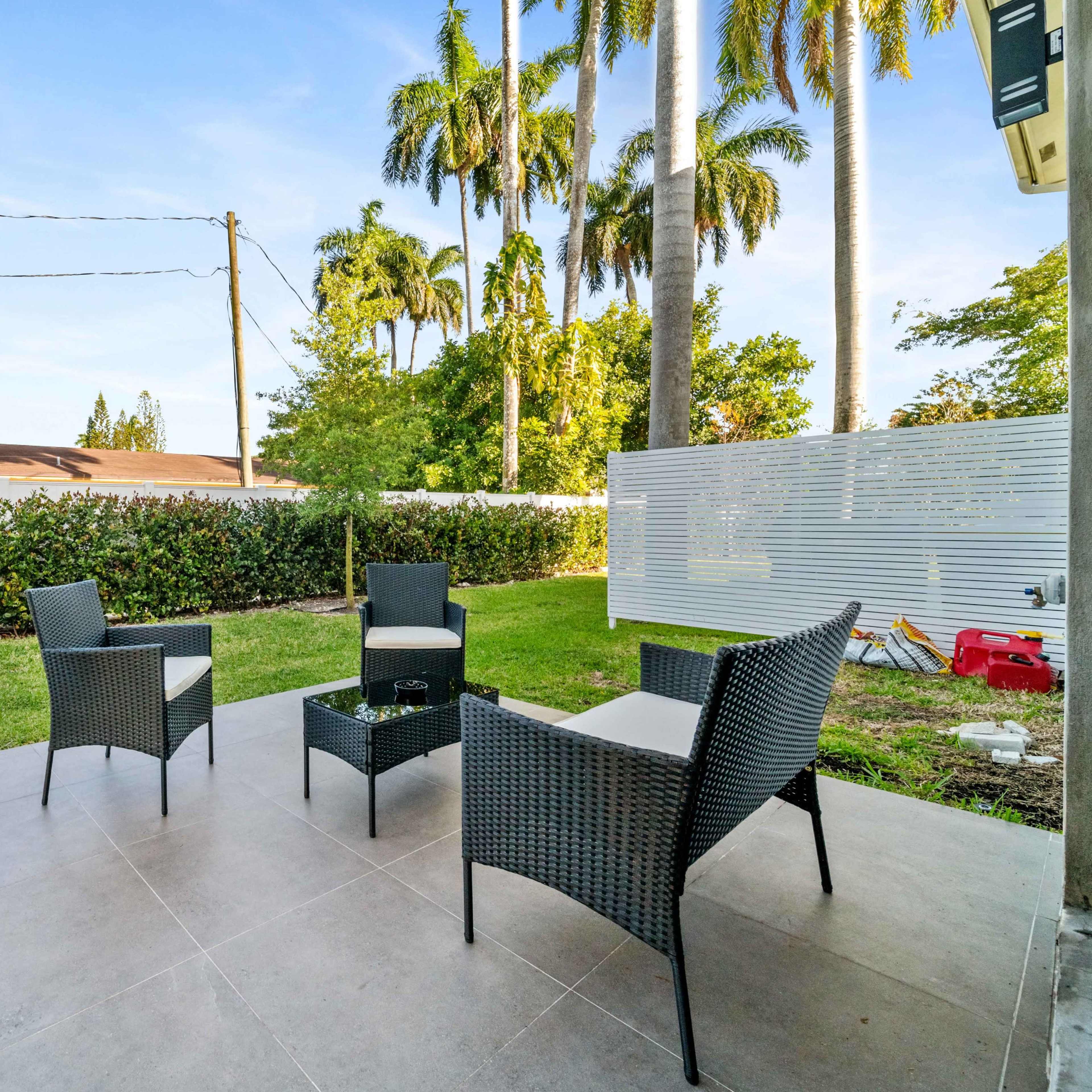 A seating area with three chairs and a small table on a tiled patio surrounded by grass and tall palm trees.