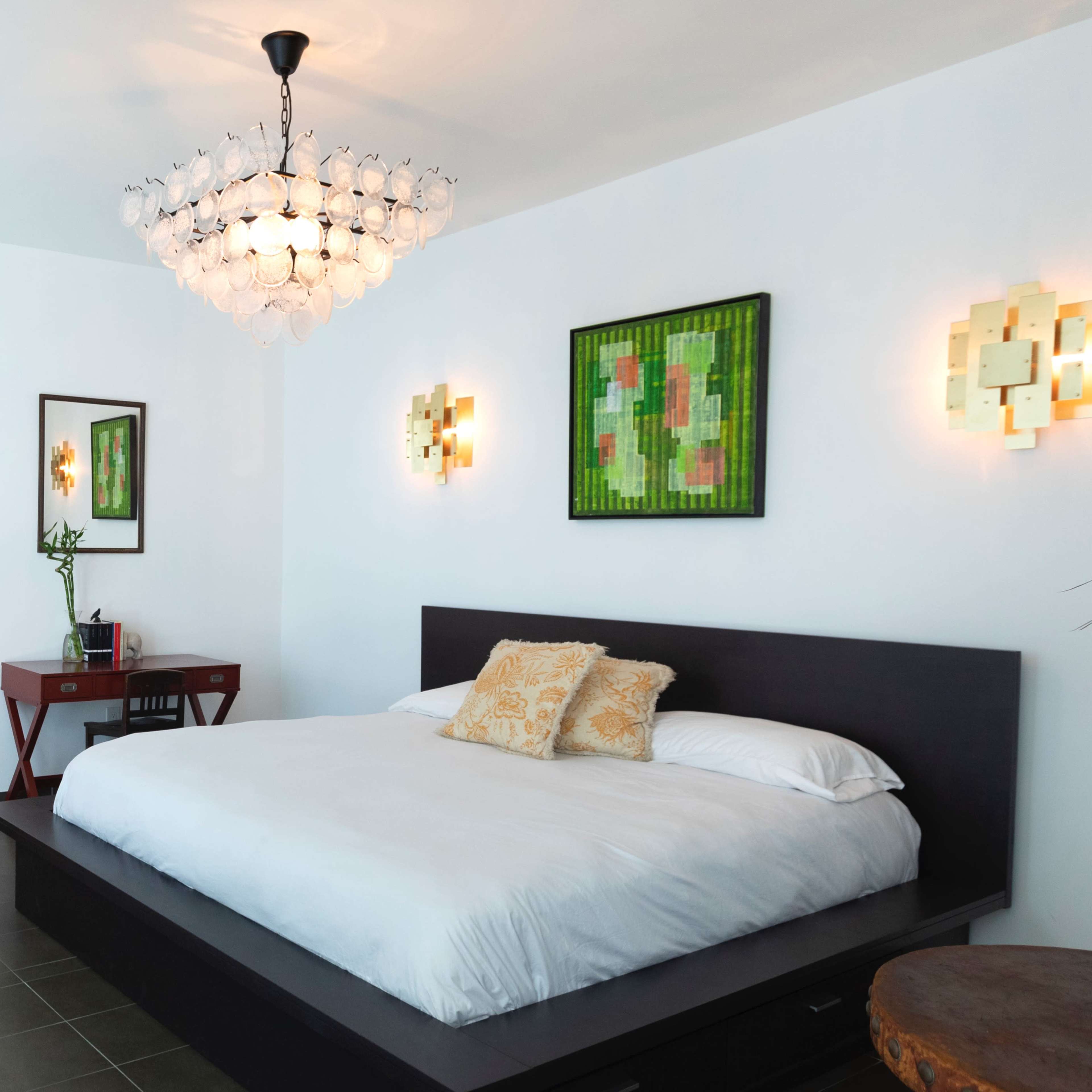 A modern bedroom features a large bed with white bedding, a decorative chandelier, two wall sconces, and abstract paintings above the bed, accompanied by a small desk and a plant in the corner.