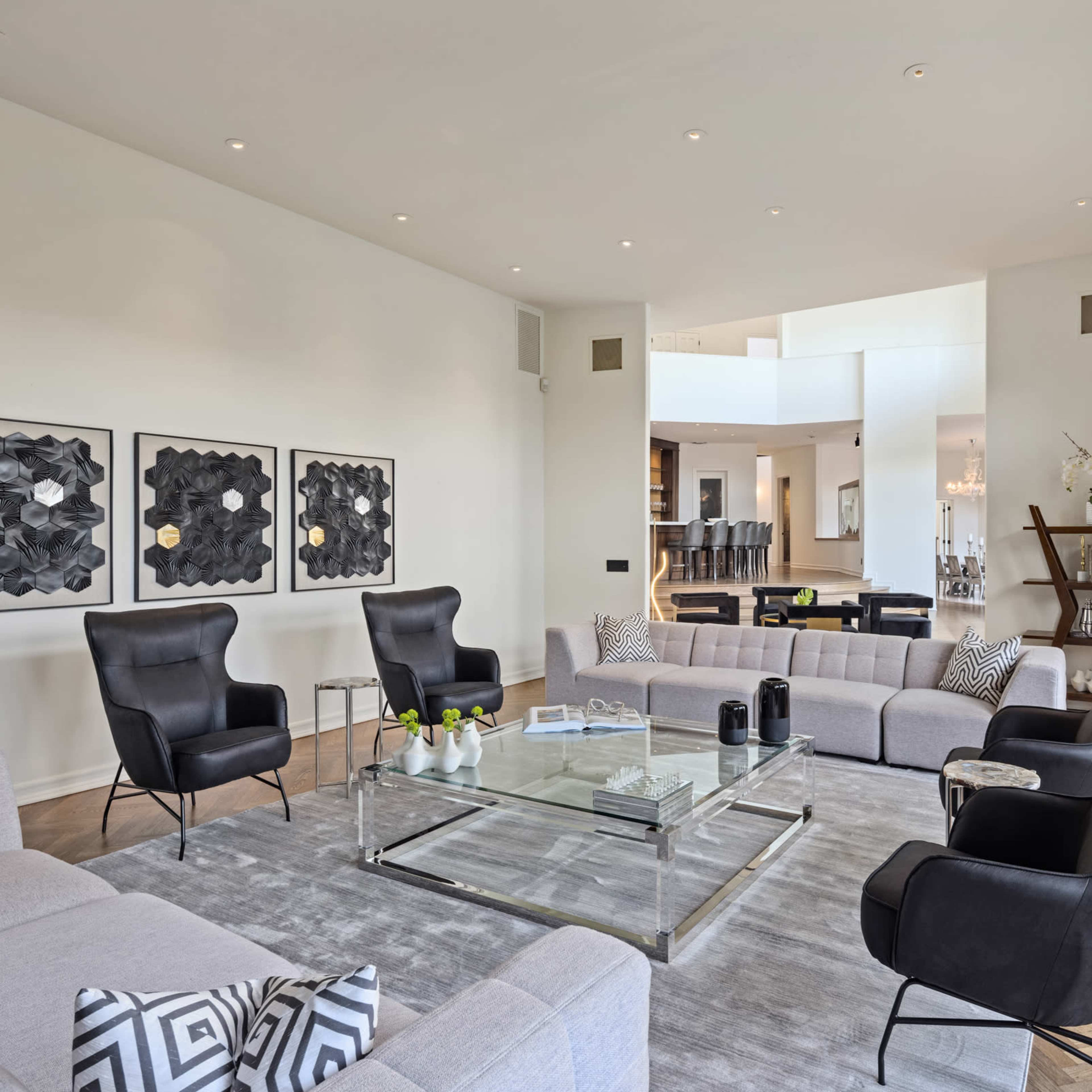 A modern living room features a large sectional sofa, two black armchairs, a glass coffee table, and wall art, all arranged in a bright space with large windows and an open layout to another room.
