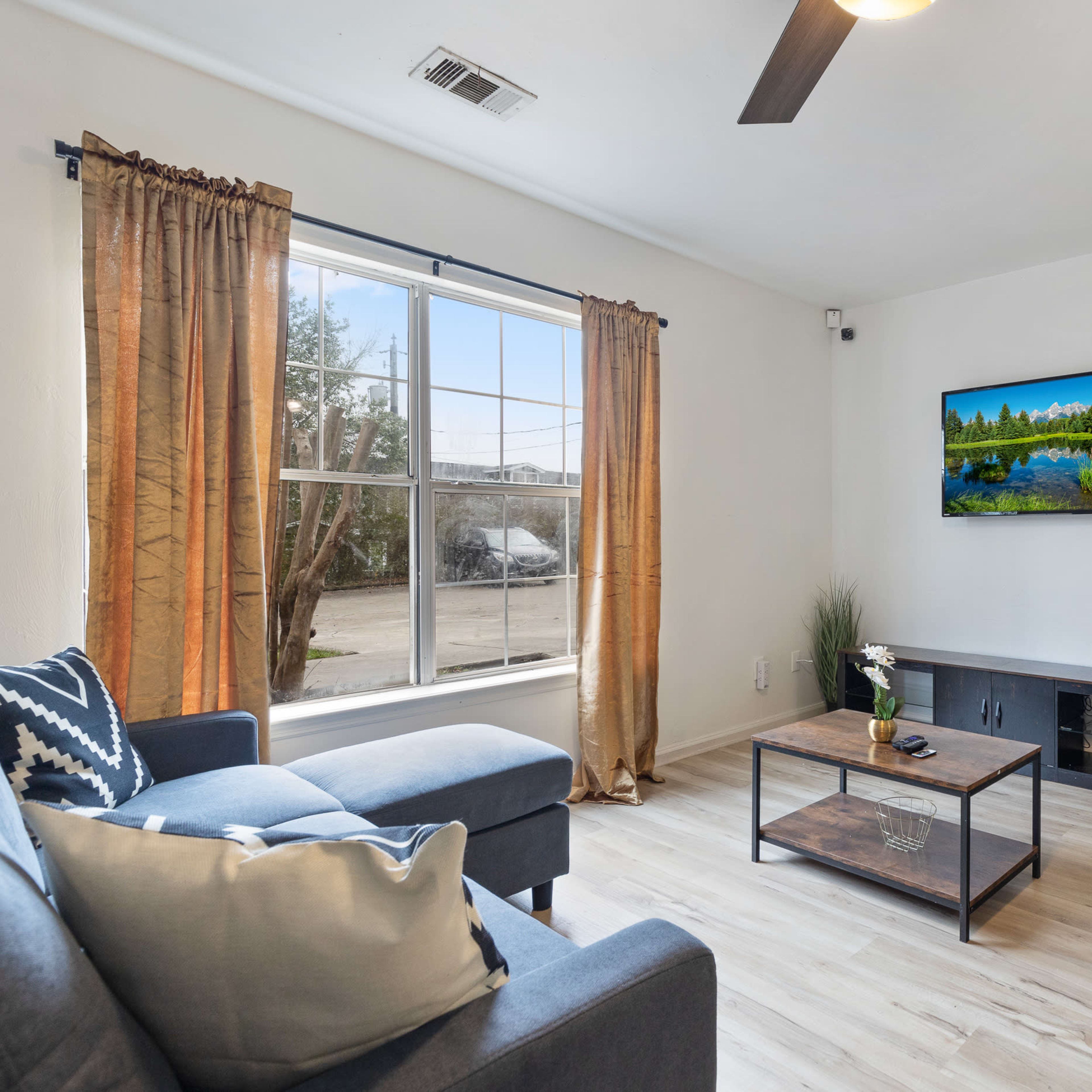 The living room features two gray sofas, a coffee table, a television mounted on the wall, and large windows with light curtains.