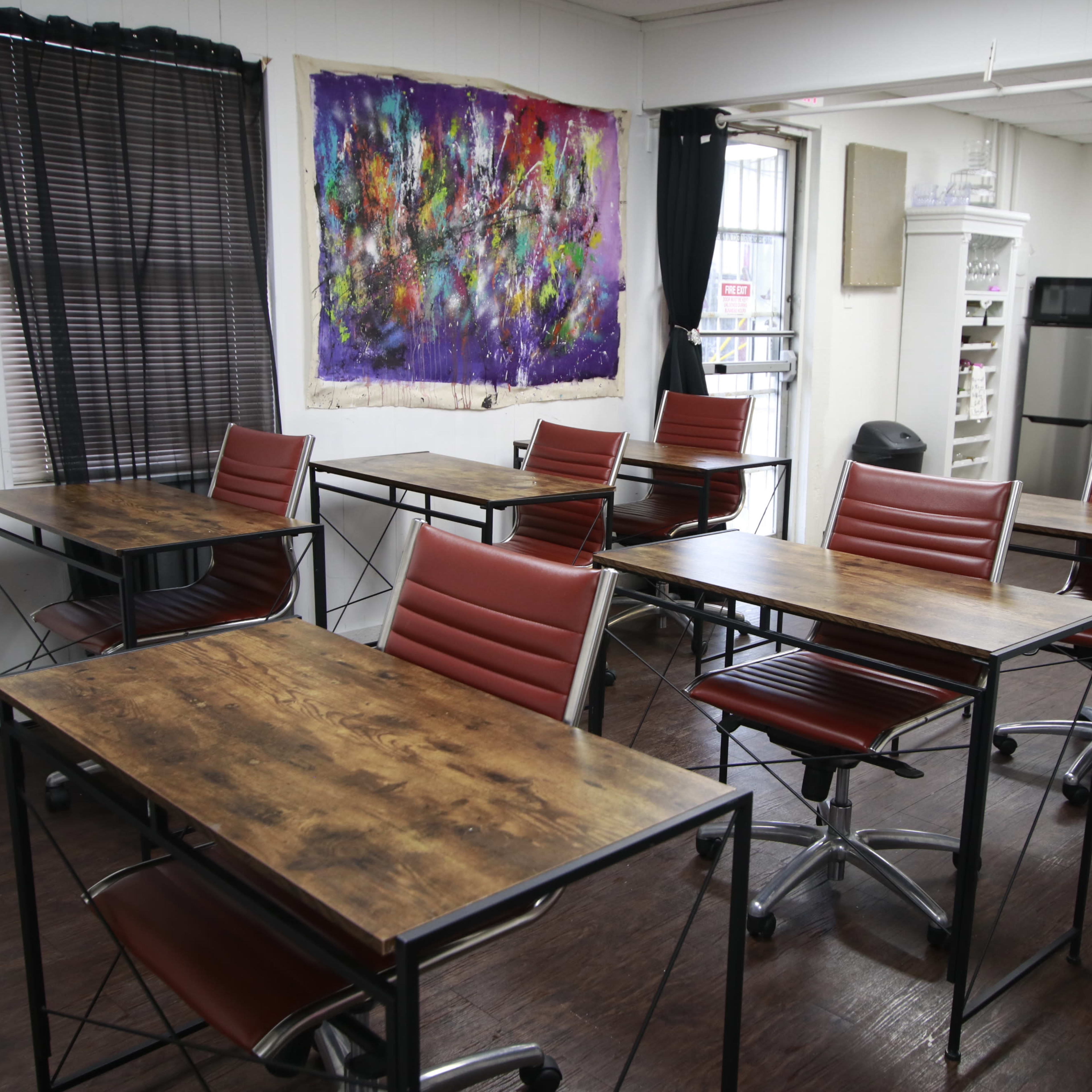 A classroom setup with multiple wooden tables and red chairs, flanked by black curtains and a colorful artwork on the wall.