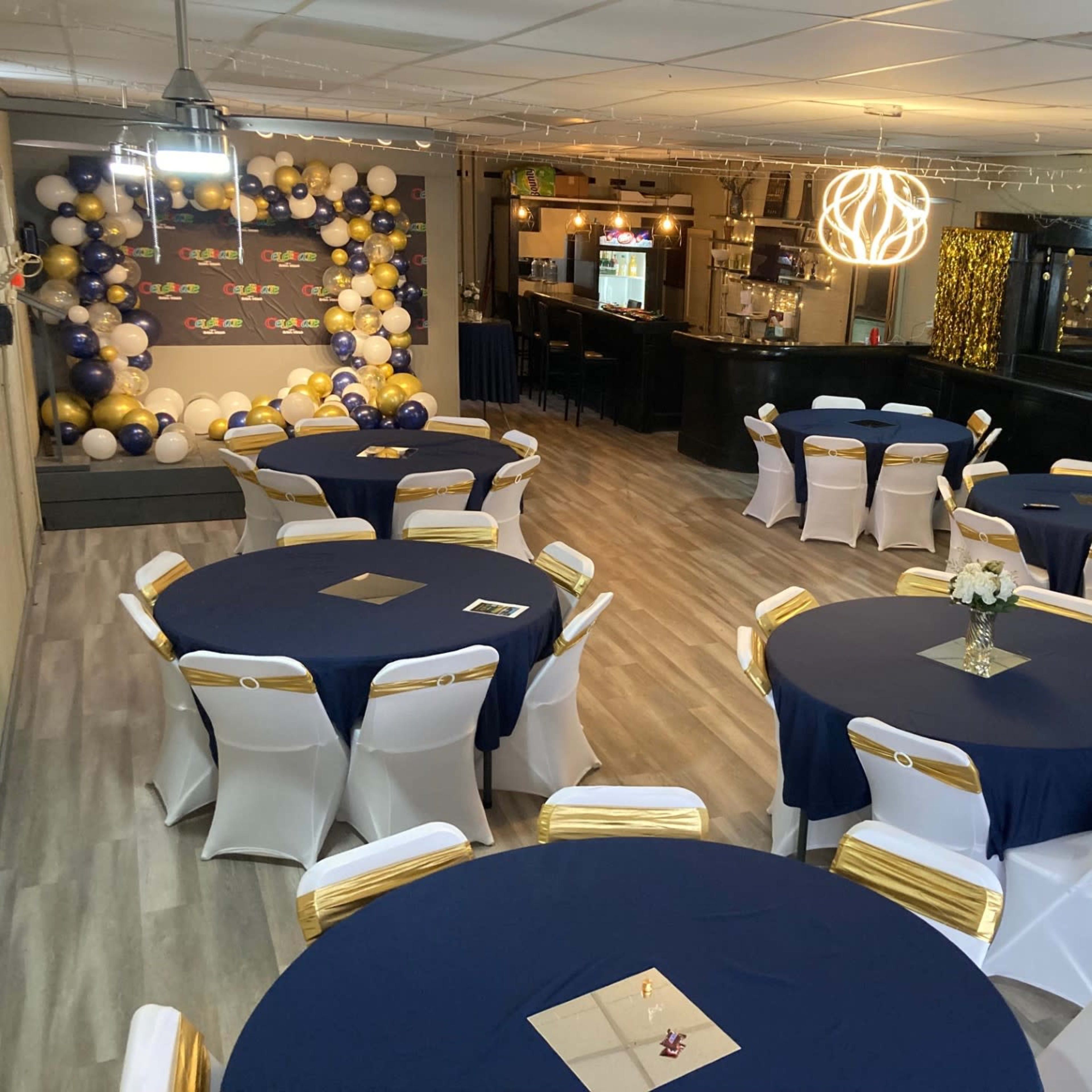 A decorated event space with round tables covered in navy blue cloths and white chairs featuring gold sashes, alongside a wall adorned with balloons.