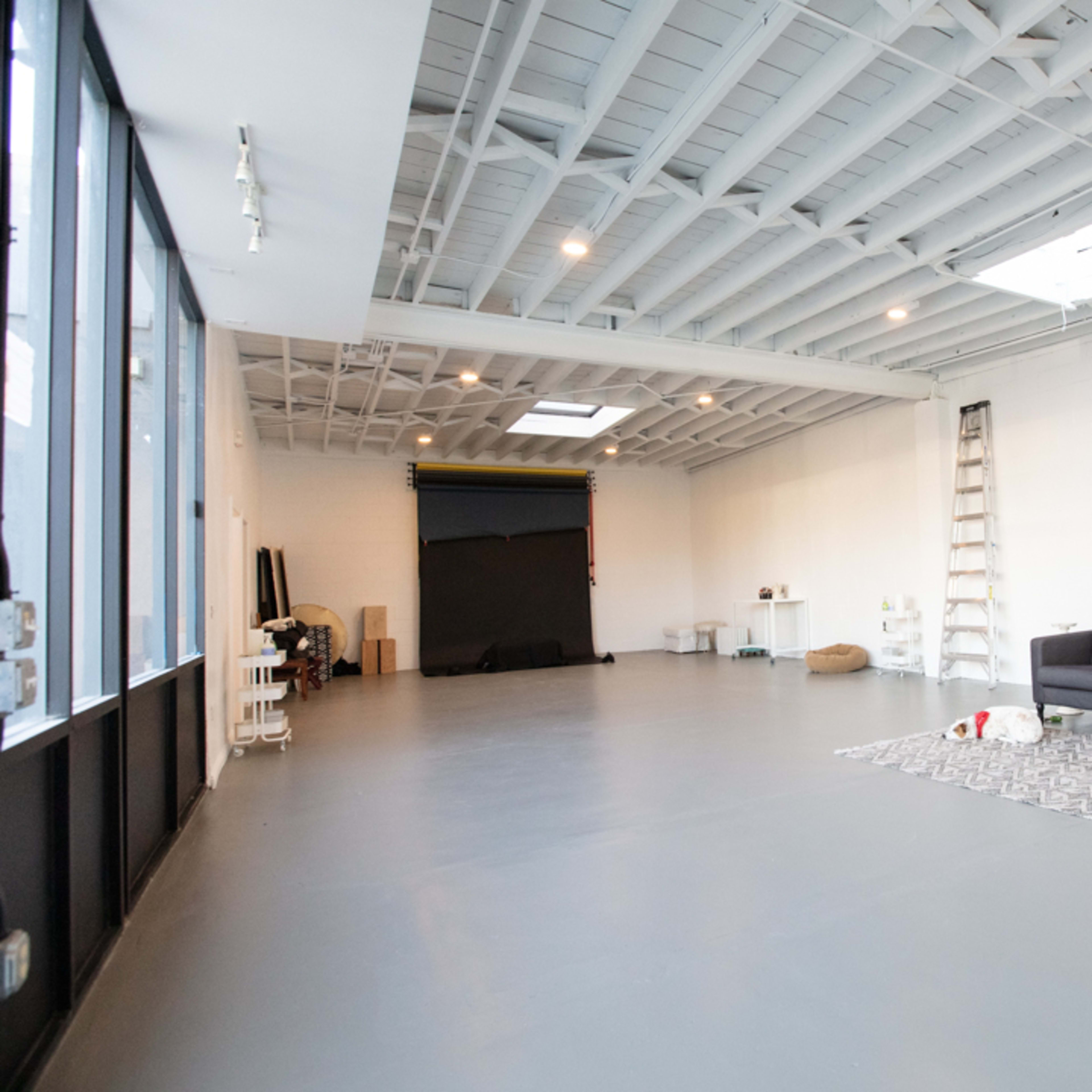 A spacious, empty studio with a gray floor, white walls, a sofa, and a large window allowing natural light to enter.