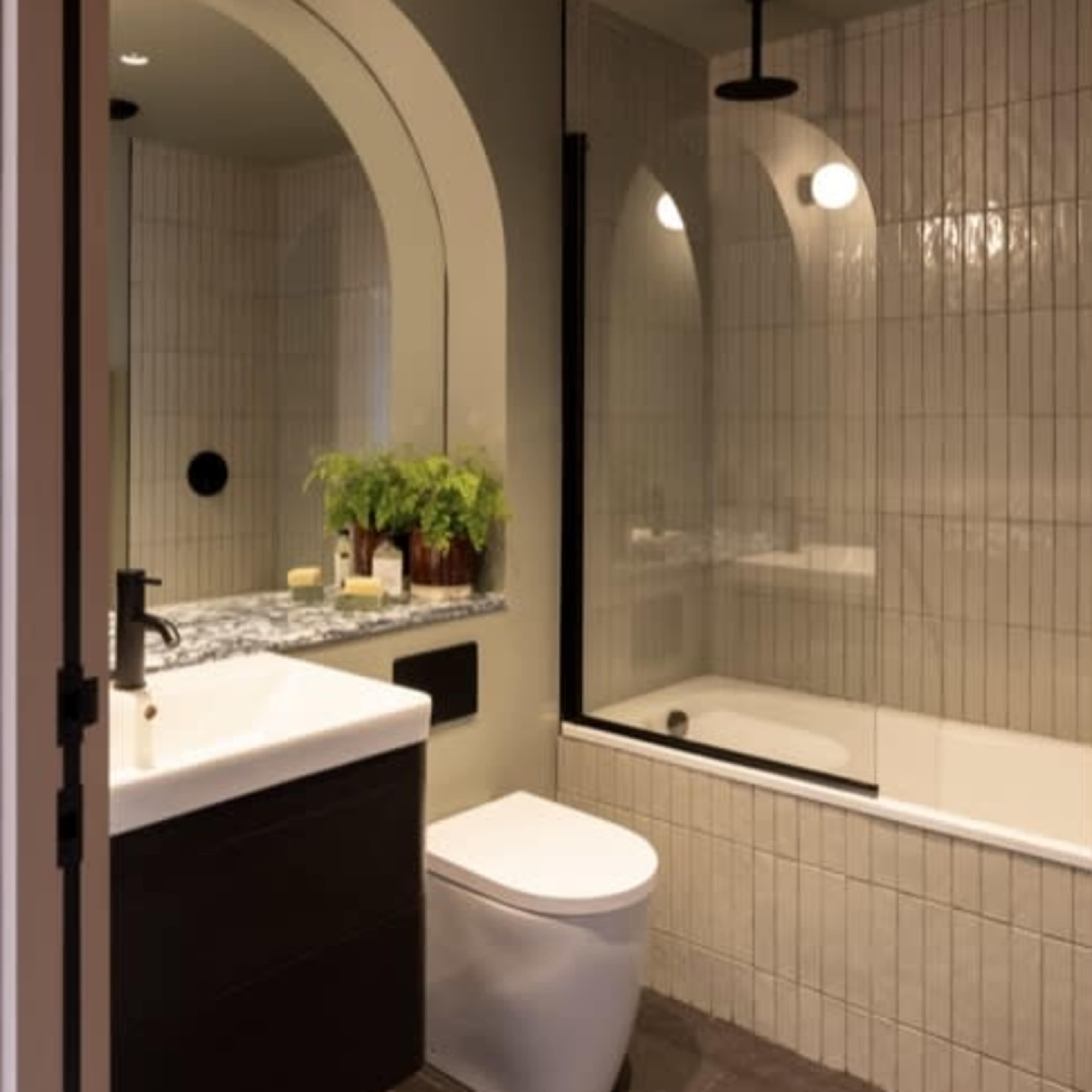 A modern bathroom featuring a bathtub with a glass shower enclosure, a wall-mounted sink with a dark cabinet, and an arched mirror, all set against vertically tiled walls.