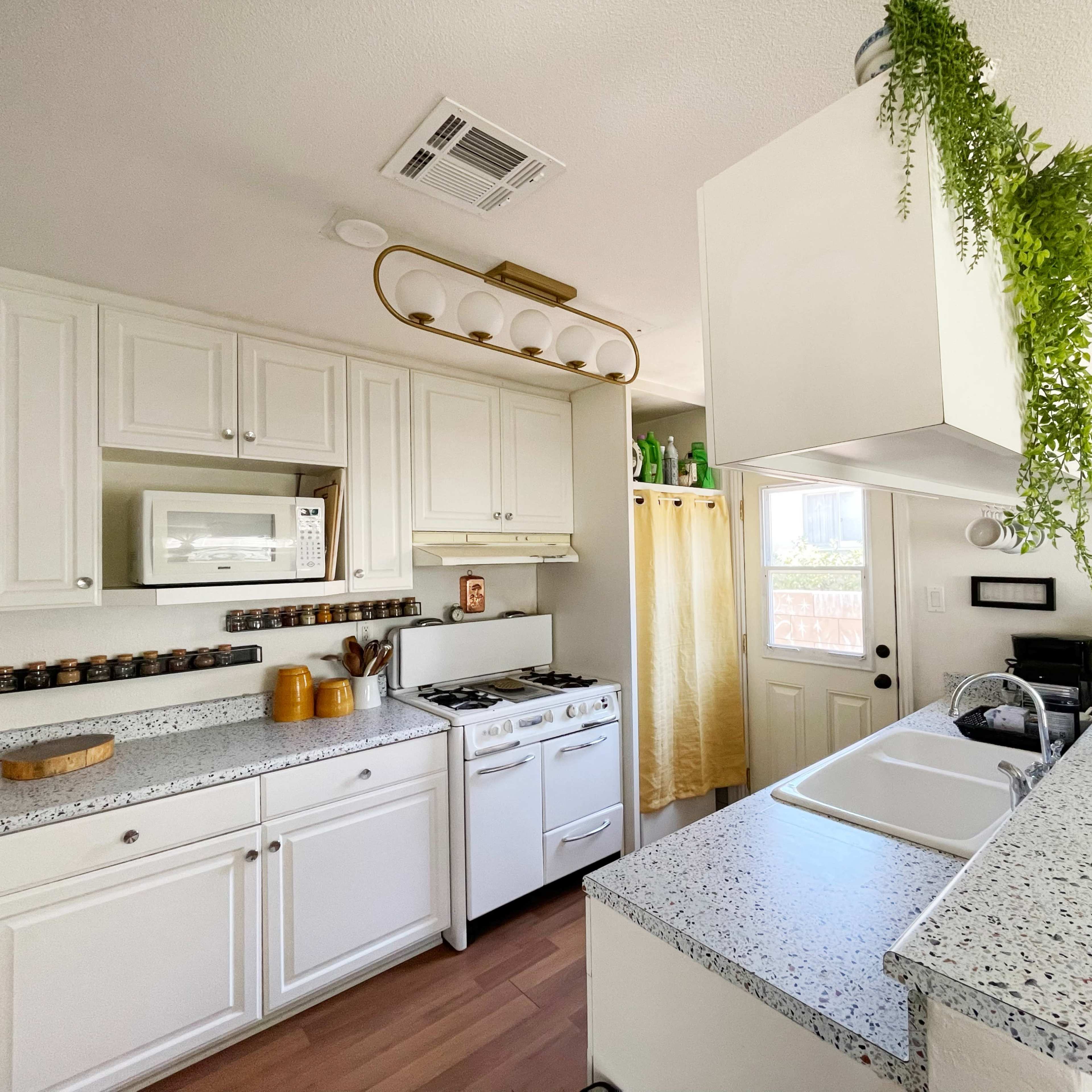 A bright kitchen features white cabinets, a stove, a microwave, and a sink, with a green plant hanging from the ceiling and wooden accents.