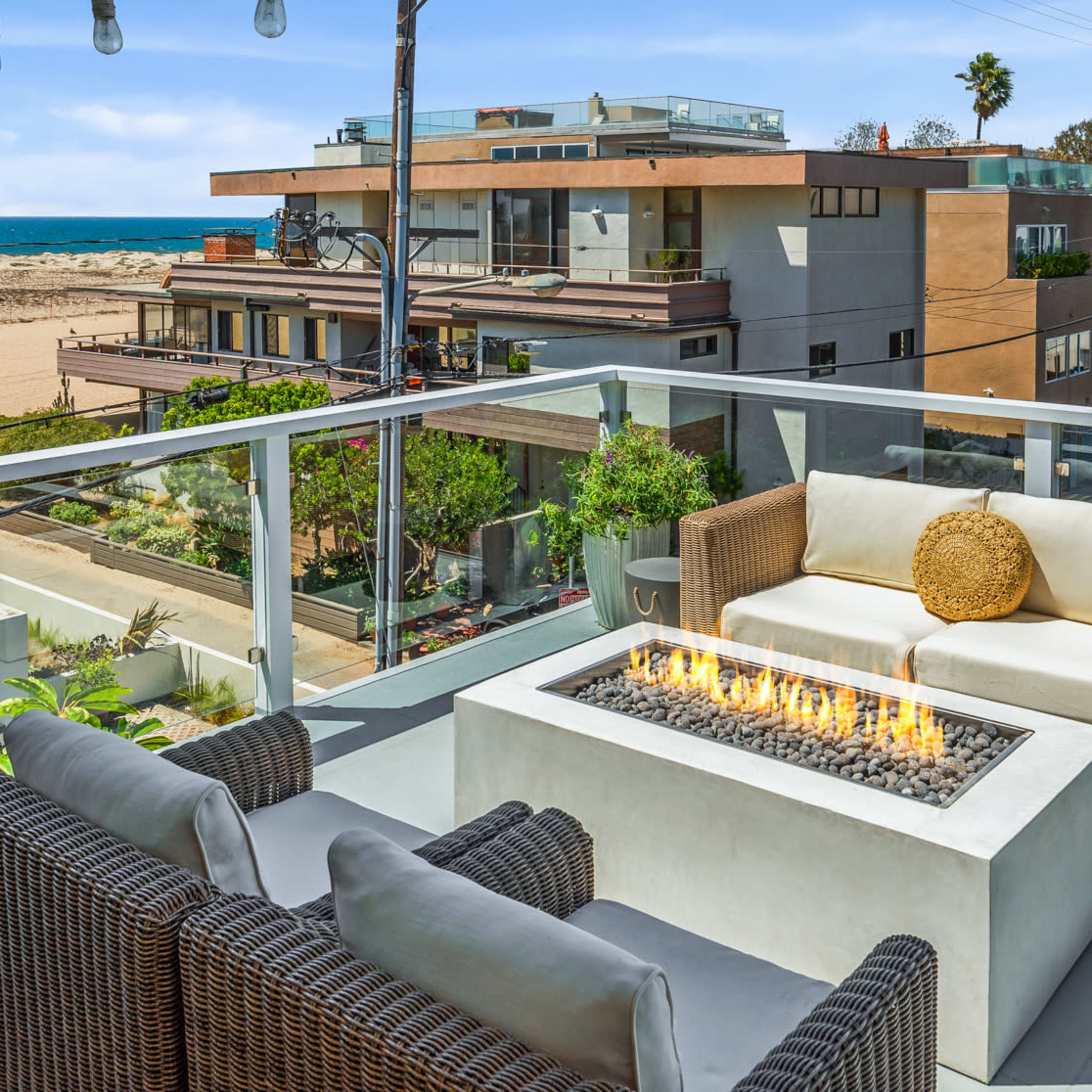 A balcony features a fire pit surrounded by outdoor seating, overlooking a sandy beach and modern buildings.