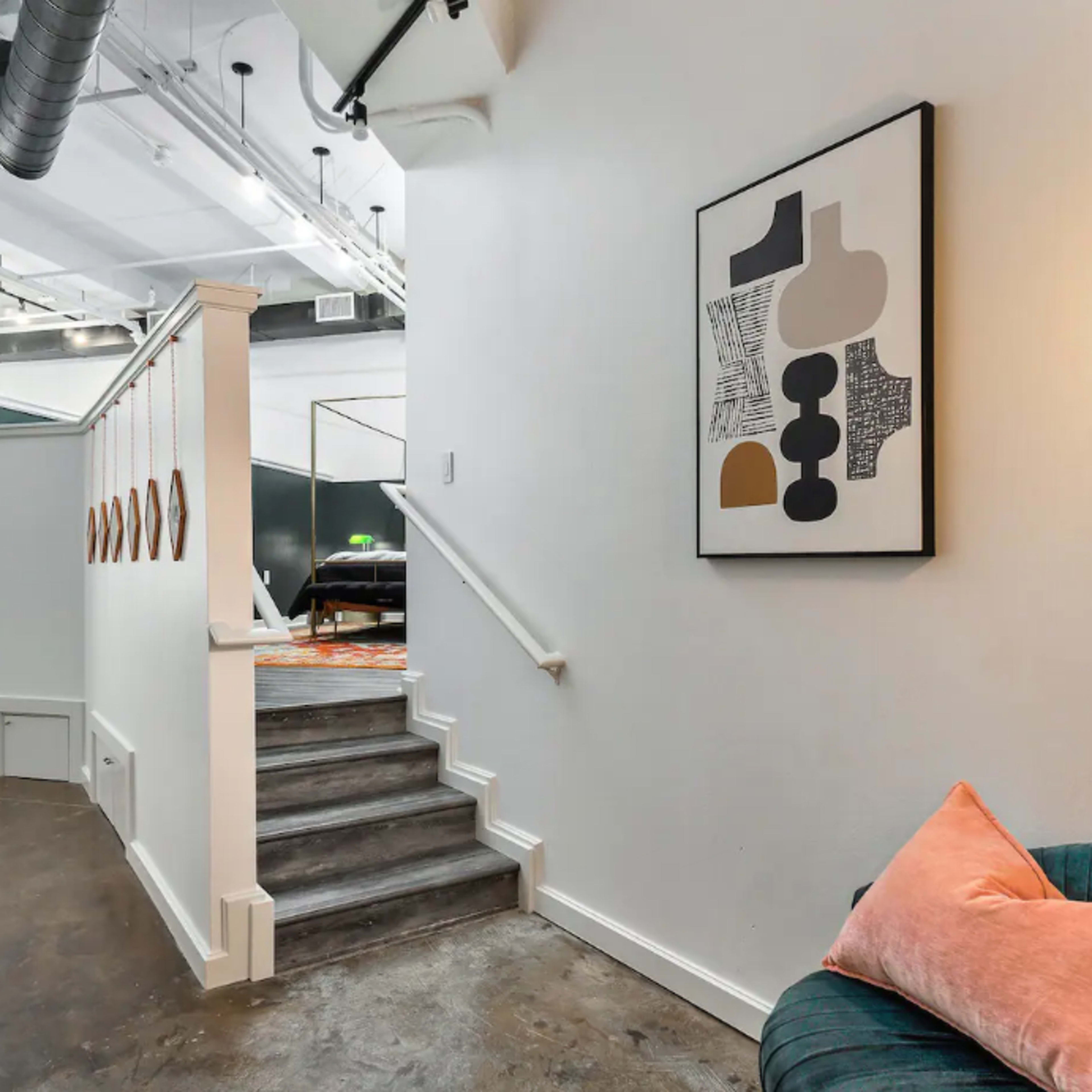 A well-lit hallway features a staircase leading to an open space, with a framed artwork on the wall and a plush chair by the side.
