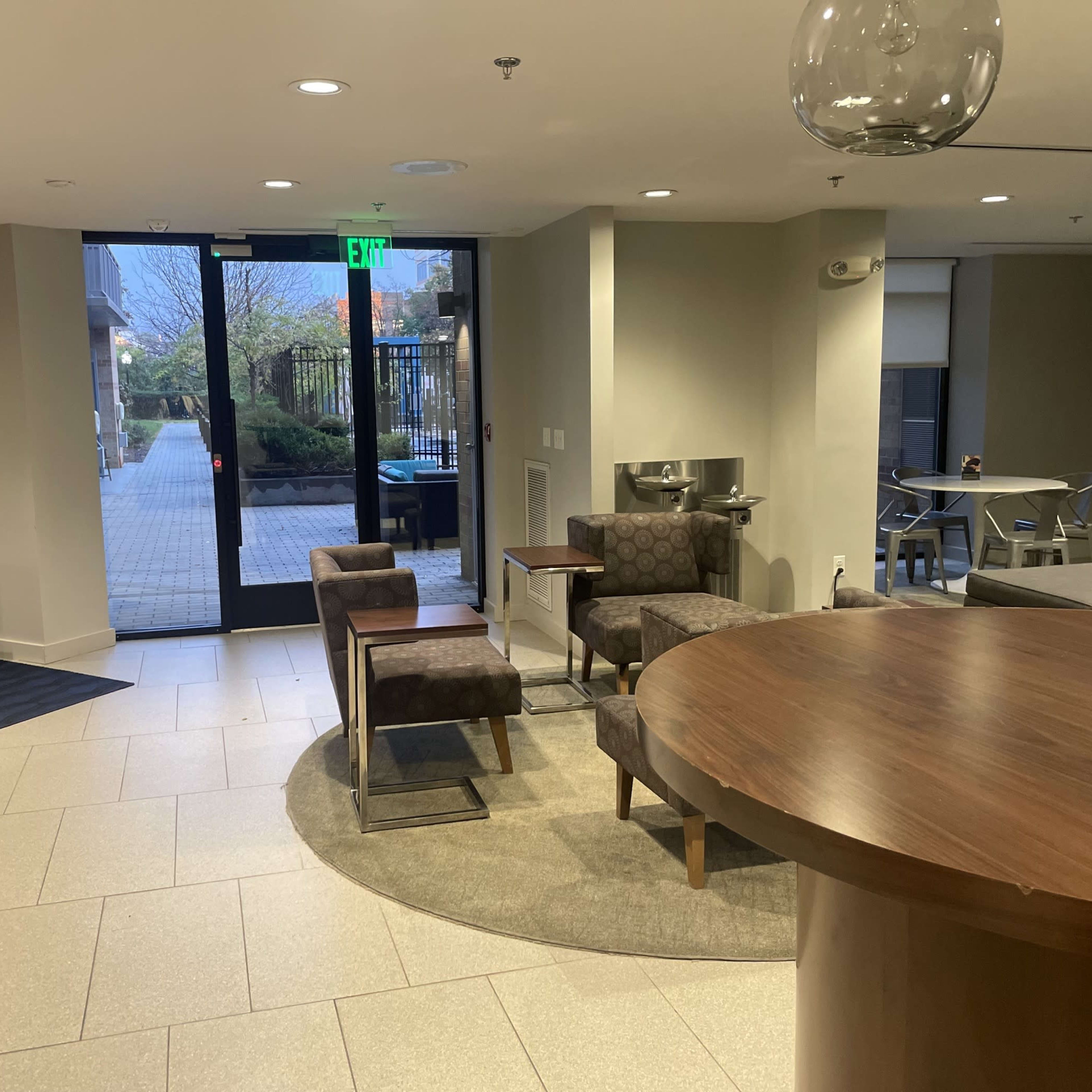 The interior of a modern lobby features a round wooden table, several chairs, and a large entrance leading to an outdoor area.