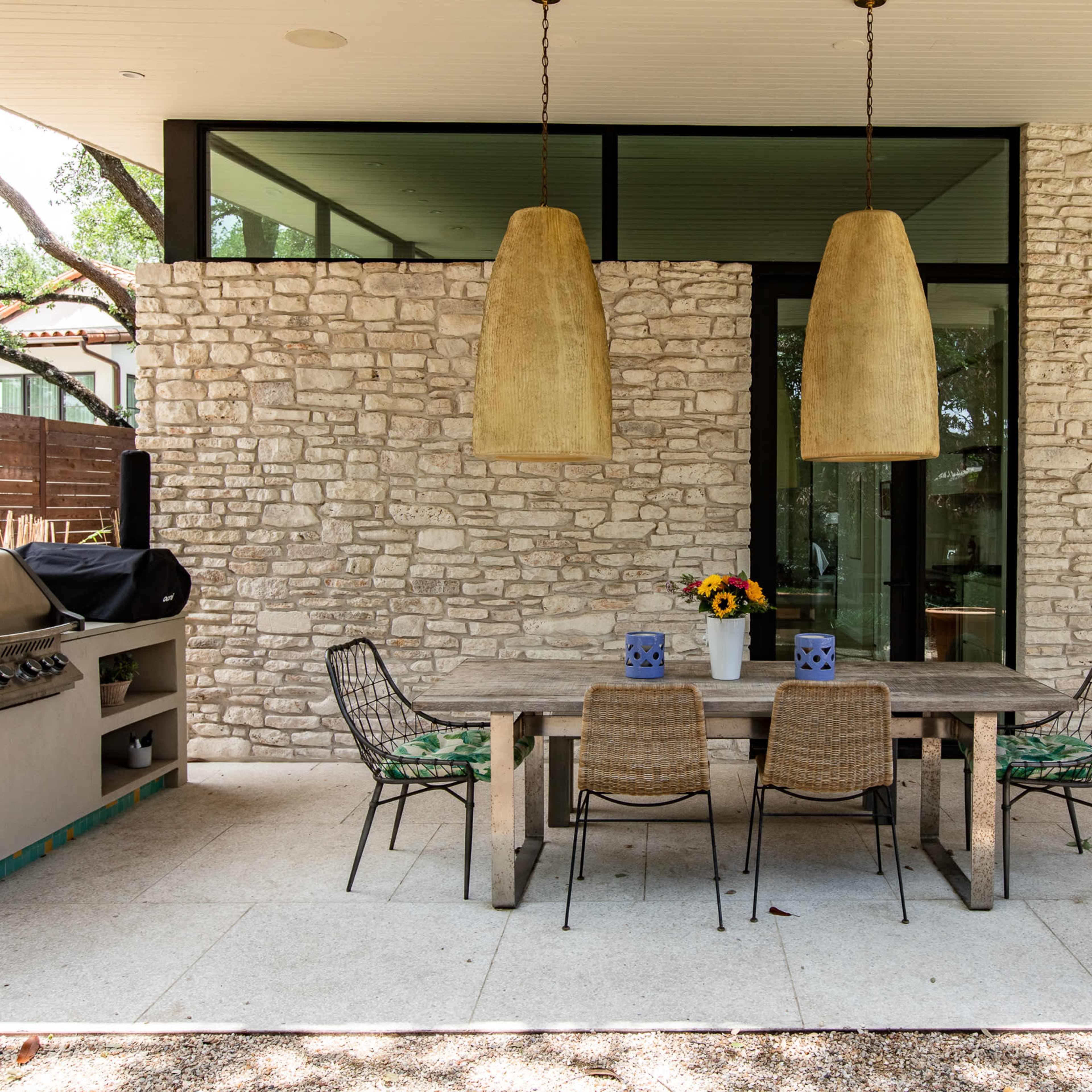 An outdoor dining area with a stone wall, a wooden table surrounded by four chairs, and two large pendant lights overhead.