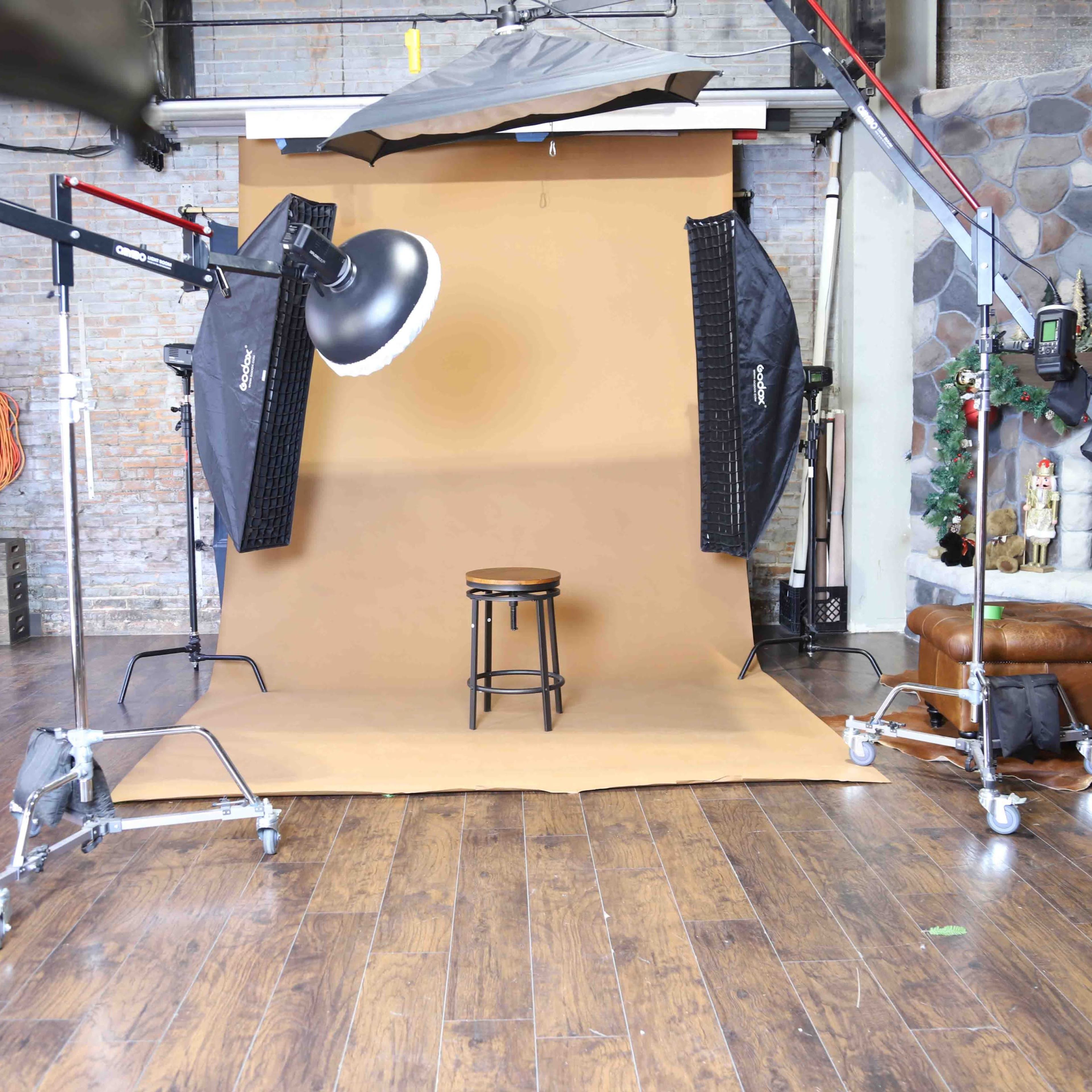 A photography studio setup featuring a brown backdrop, a stool in the center, and various lighting equipment arranged around the scene.