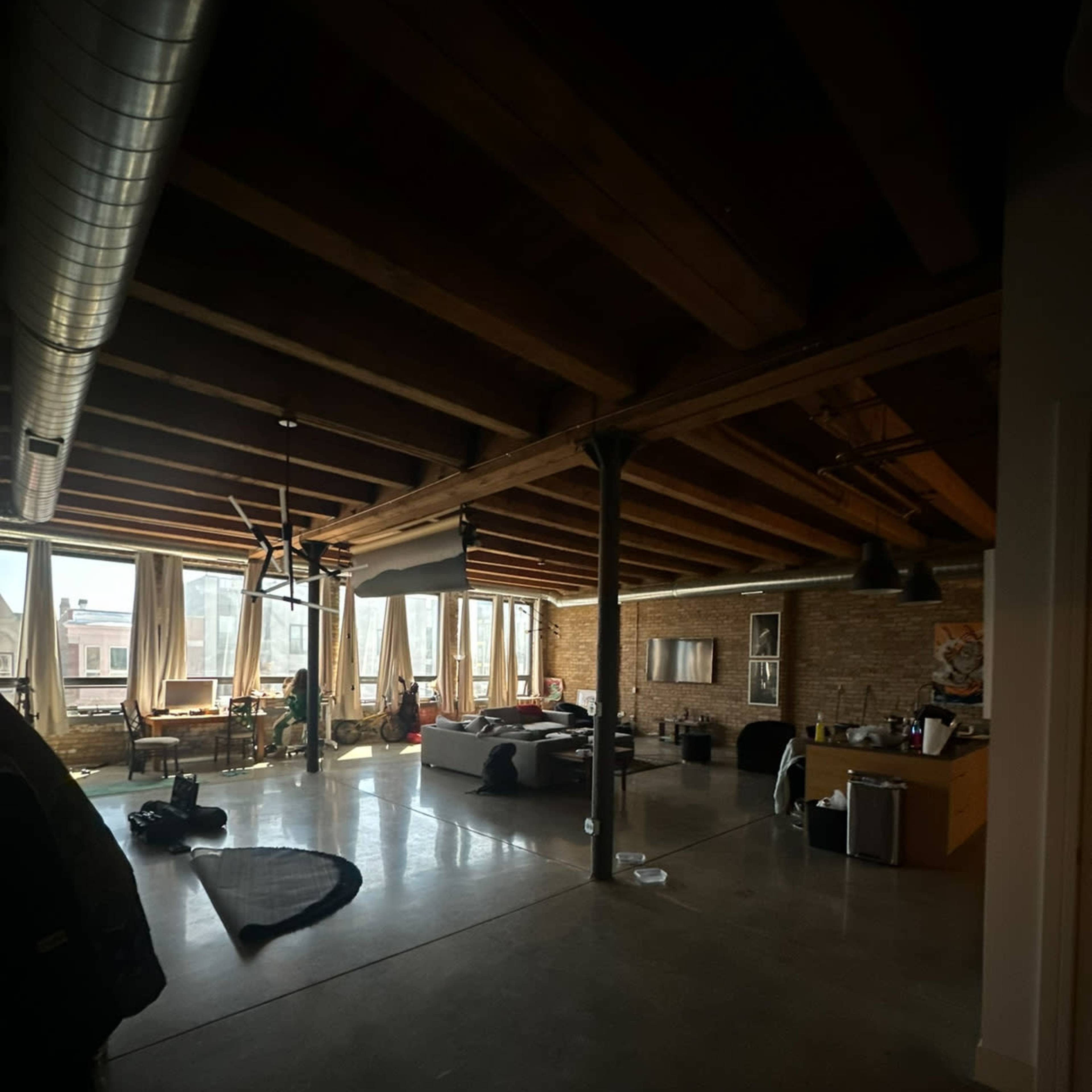 A spacious loft apartment with exposed wooden beams, large windows, and a mix of furniture scattered throughout.