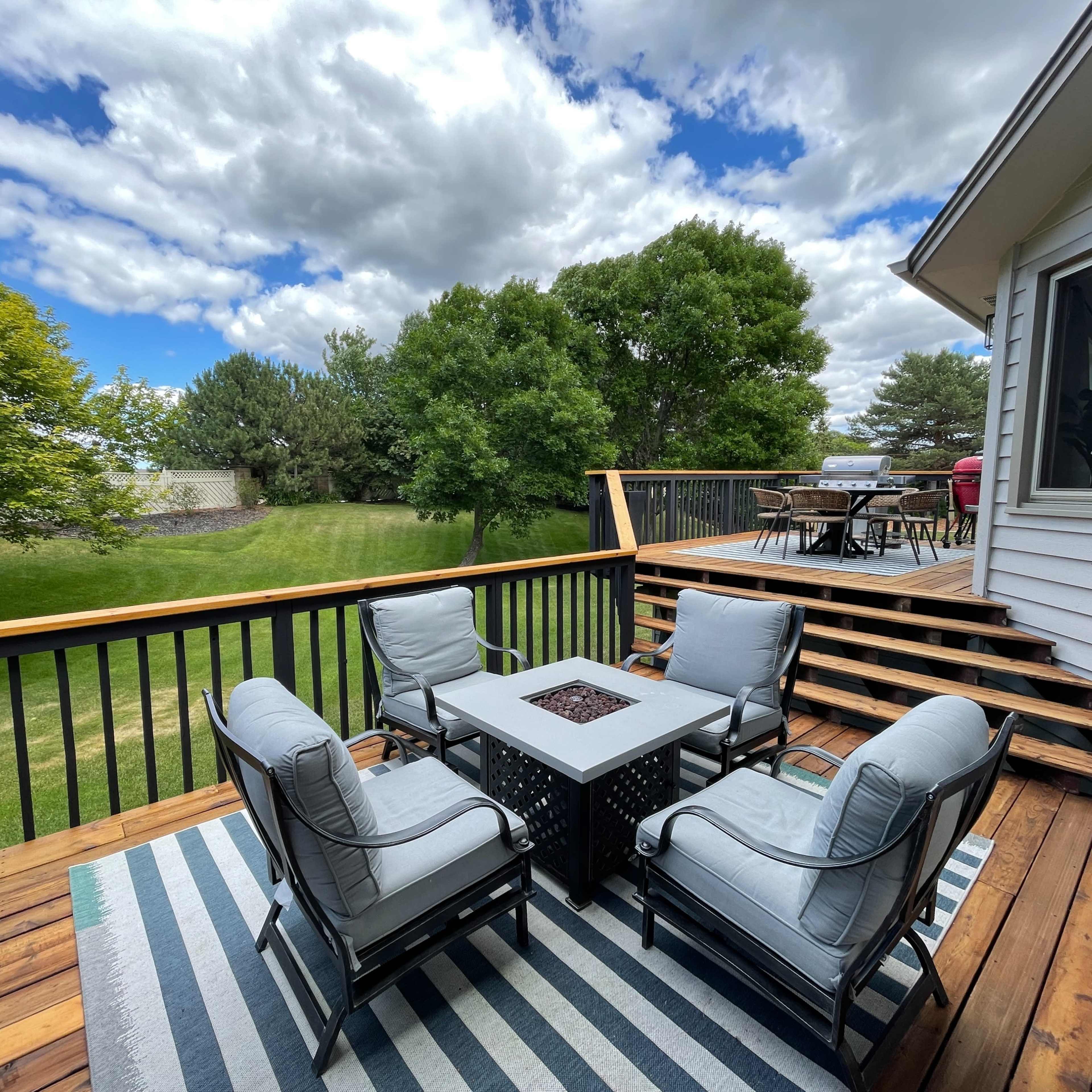 A wooden deck features a seating arrangement with four chairs and a table, surrounded by greenery and under a partly cloudy sky.