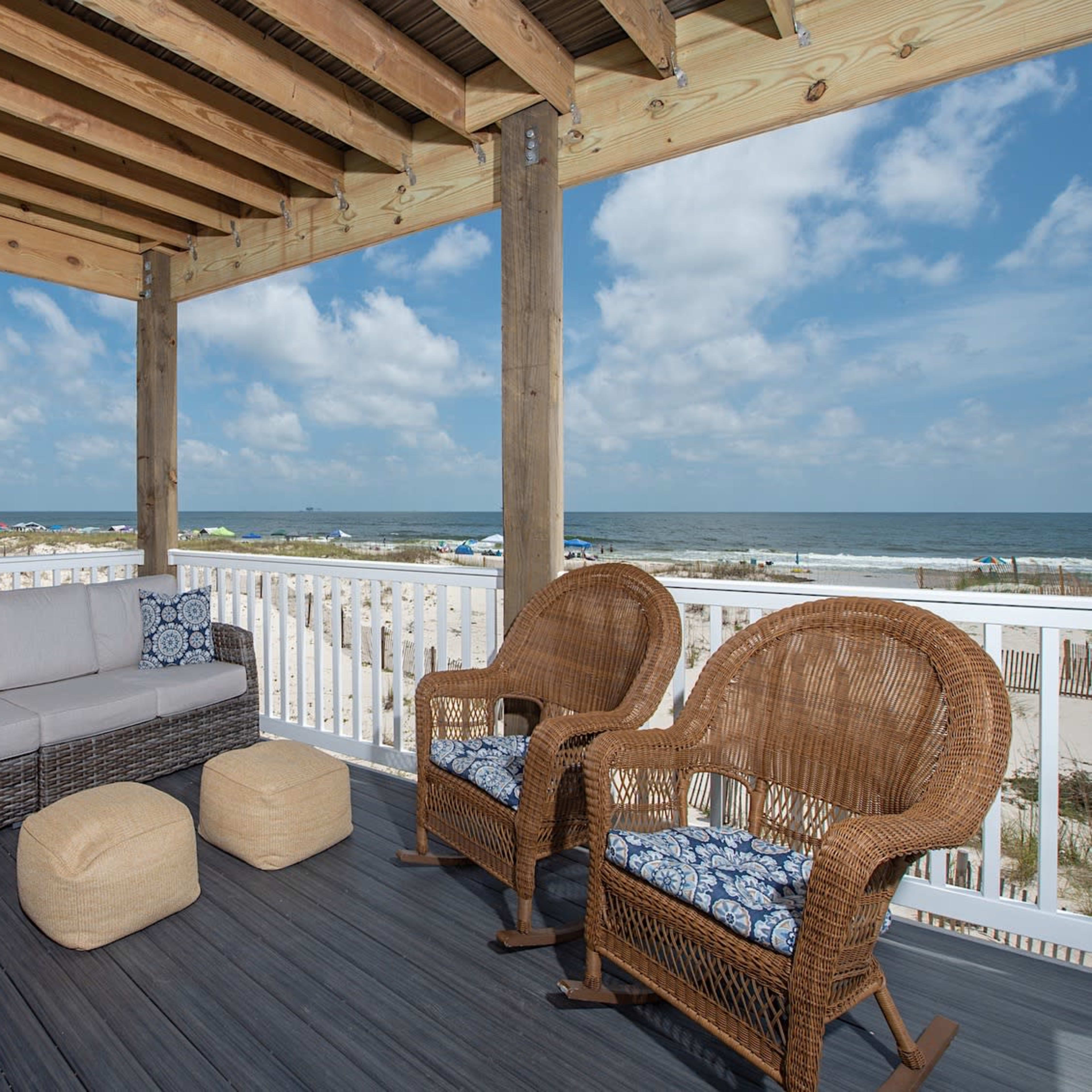 A covered porch with wicker furniture overlooks the beach and ocean under a partly cloudy sky.