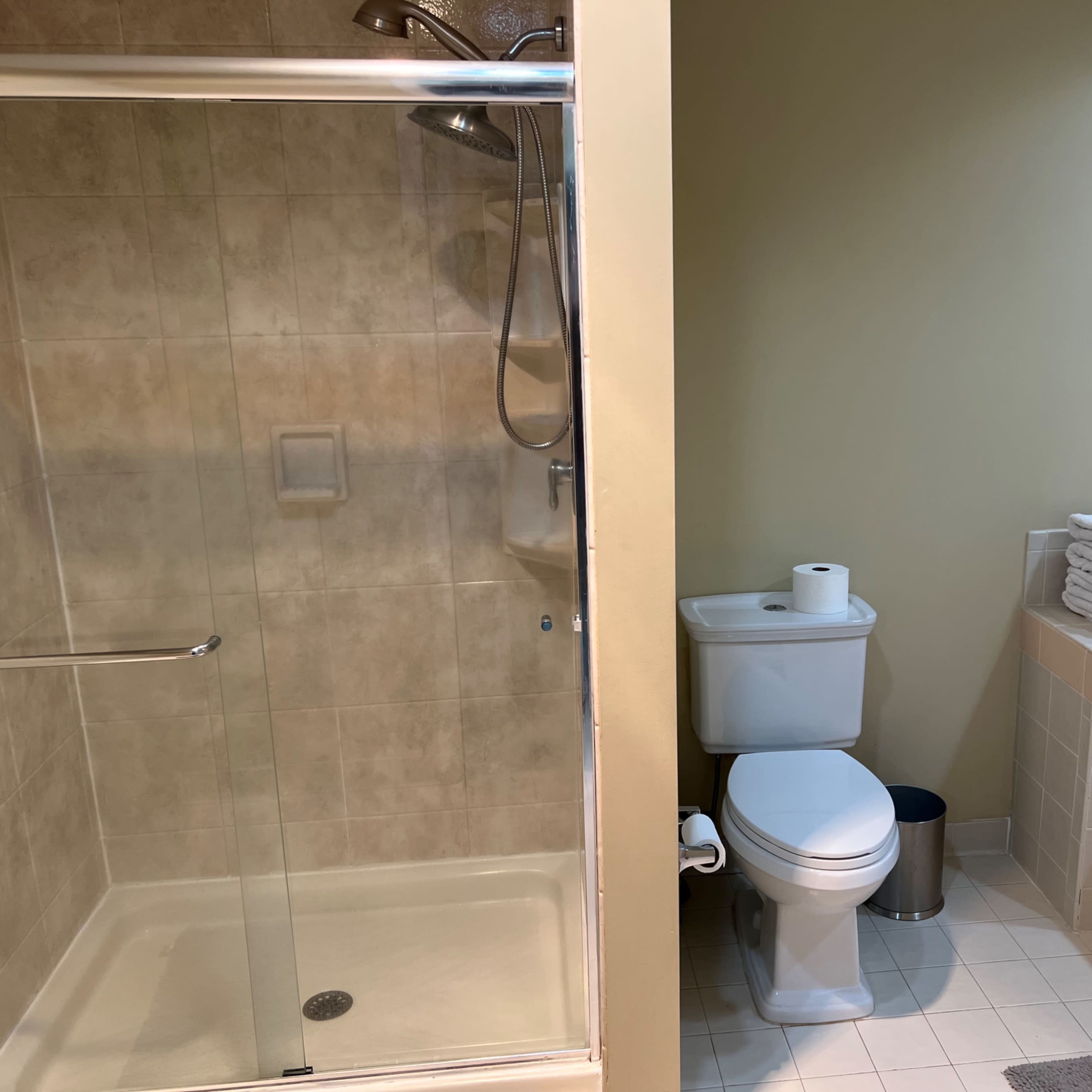 A bathroom divided into two sections, featuring a glass shower enclosure on the left and a toilet with folded towels beside a bathtub on the right.