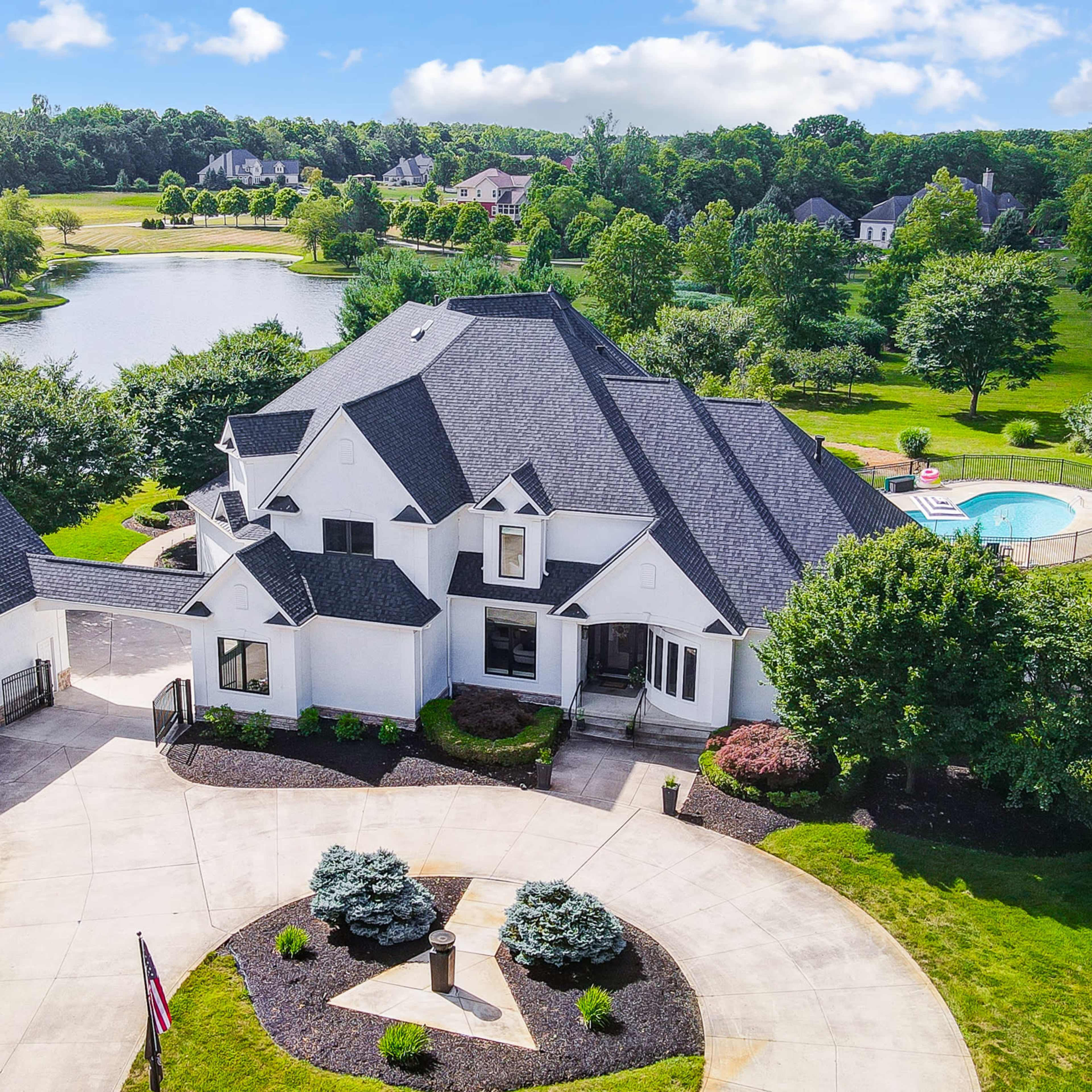 A large, modern home with a prominent roof design is situated on a landscaped property near a pond and swimming pool.