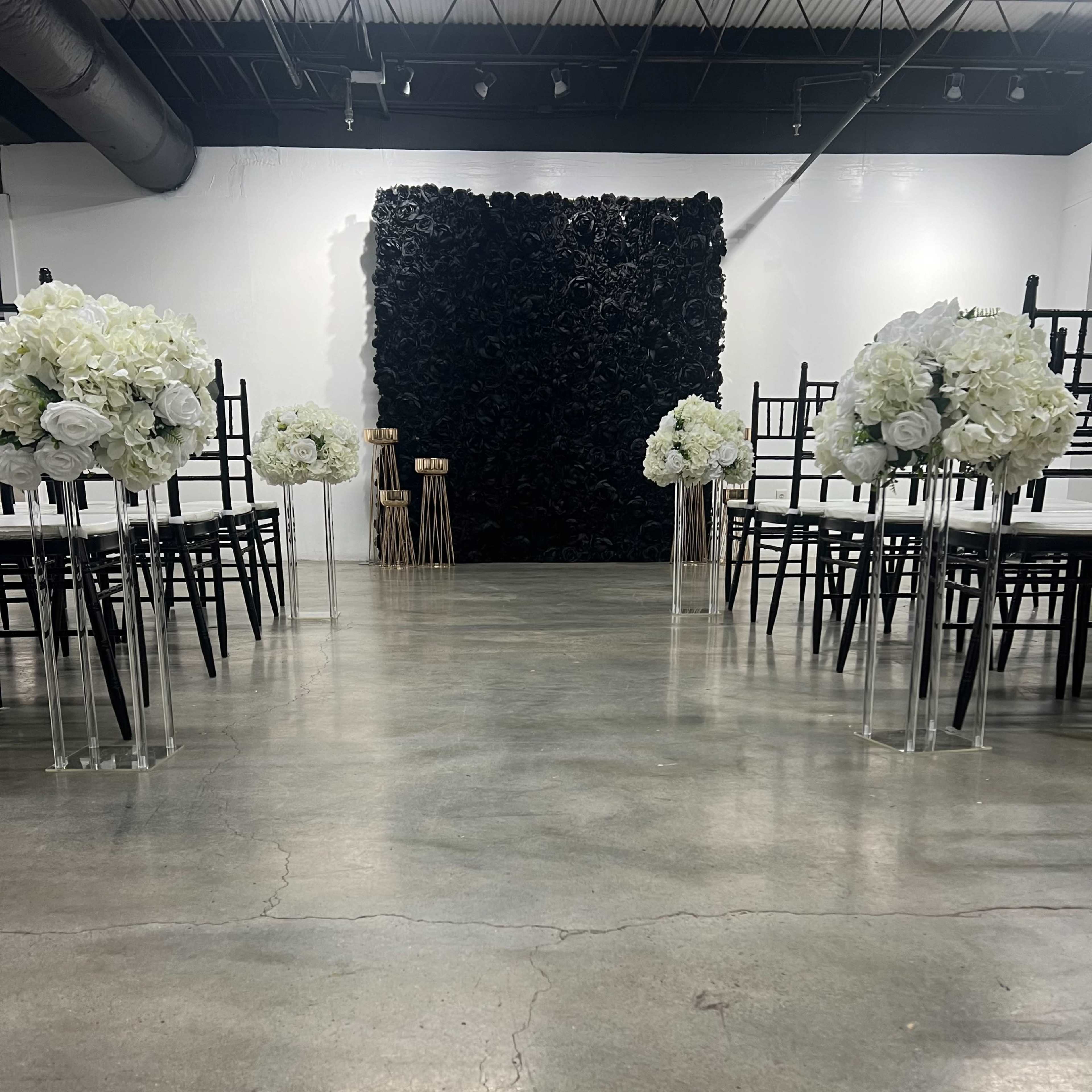 A wedding ceremony setup with rows of black chairs facing a textured black backdrop, flanked by arrangements of white flowers on clear stands.
