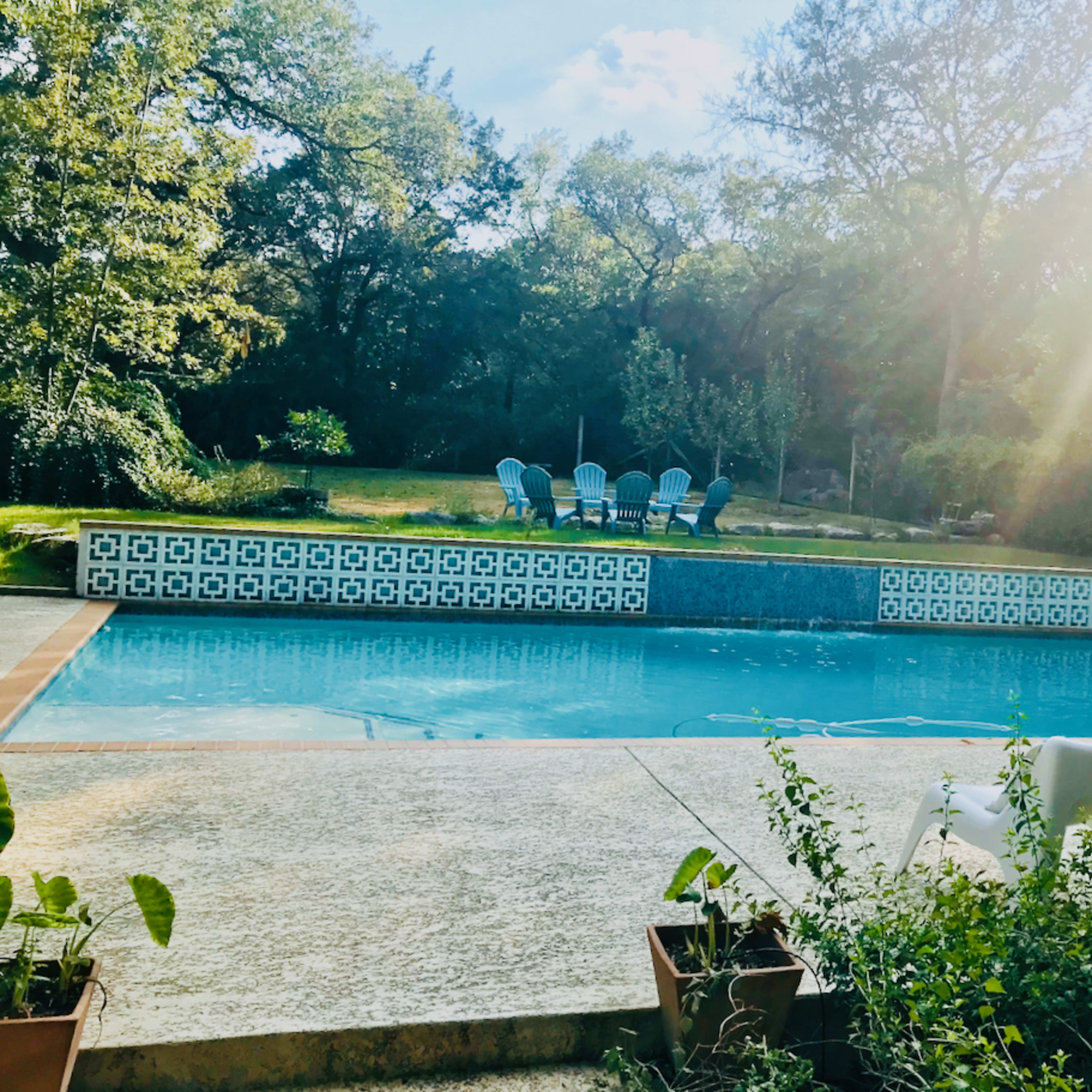 A swimming pool surrounded by greenery, with several blue chairs positioned nearby and two white lounge chairs on the pool deck.