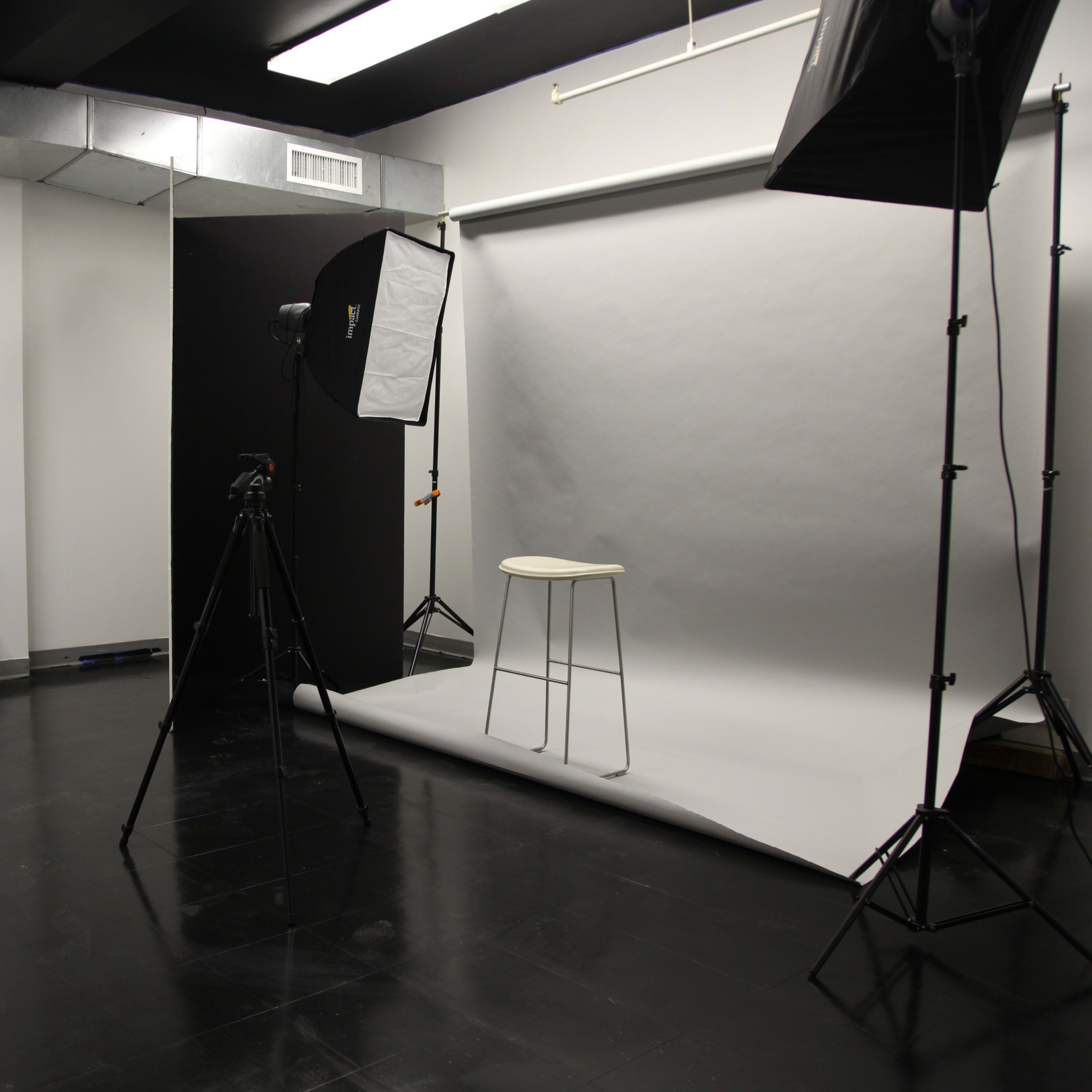 A photography studio with a gray backdrop, a stool, a camera setup on a tripod, and a mirrored vanity with a red chair.