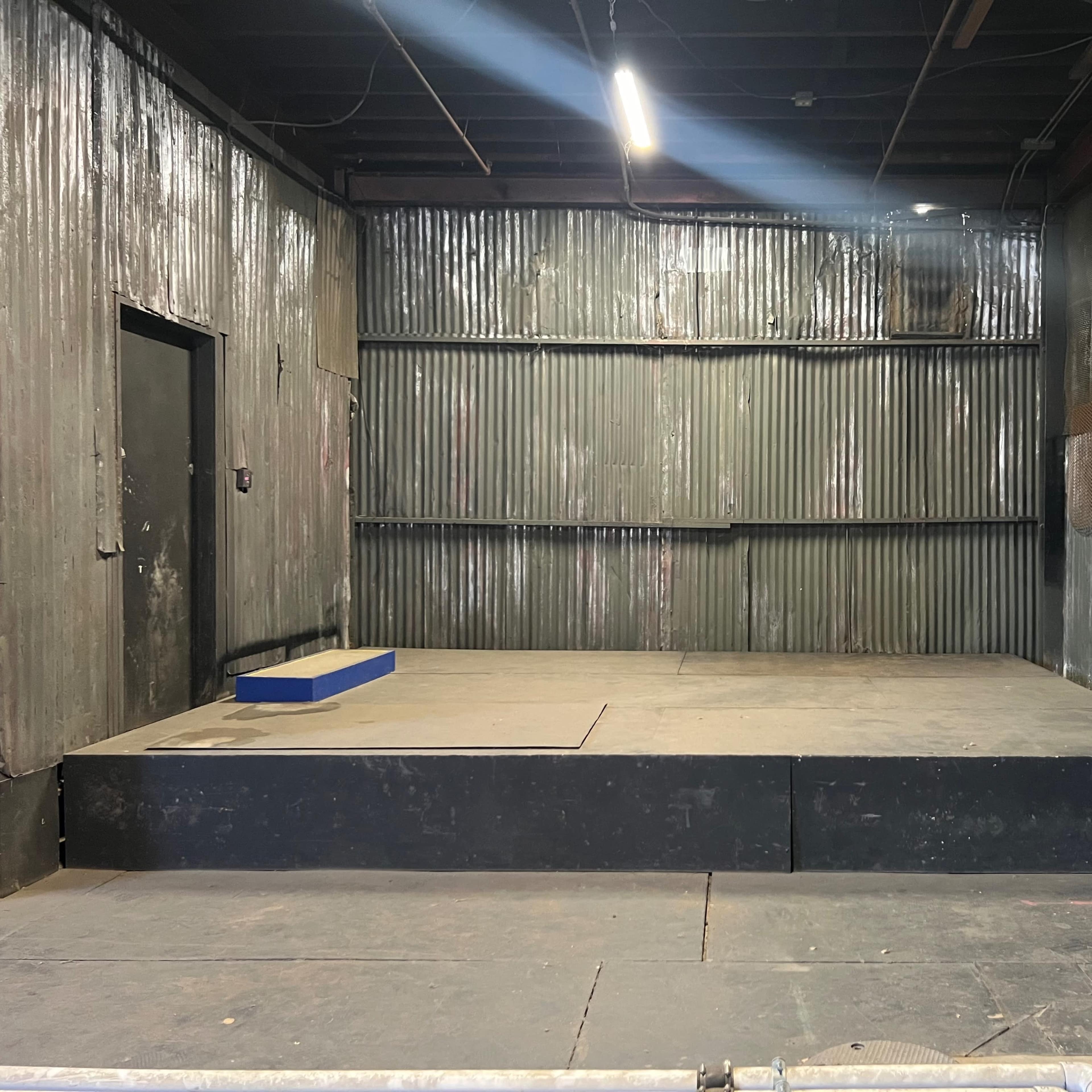 A dimly lit, empty stage with a raised platform and a dark doorway in a room with corrugated metal walls.