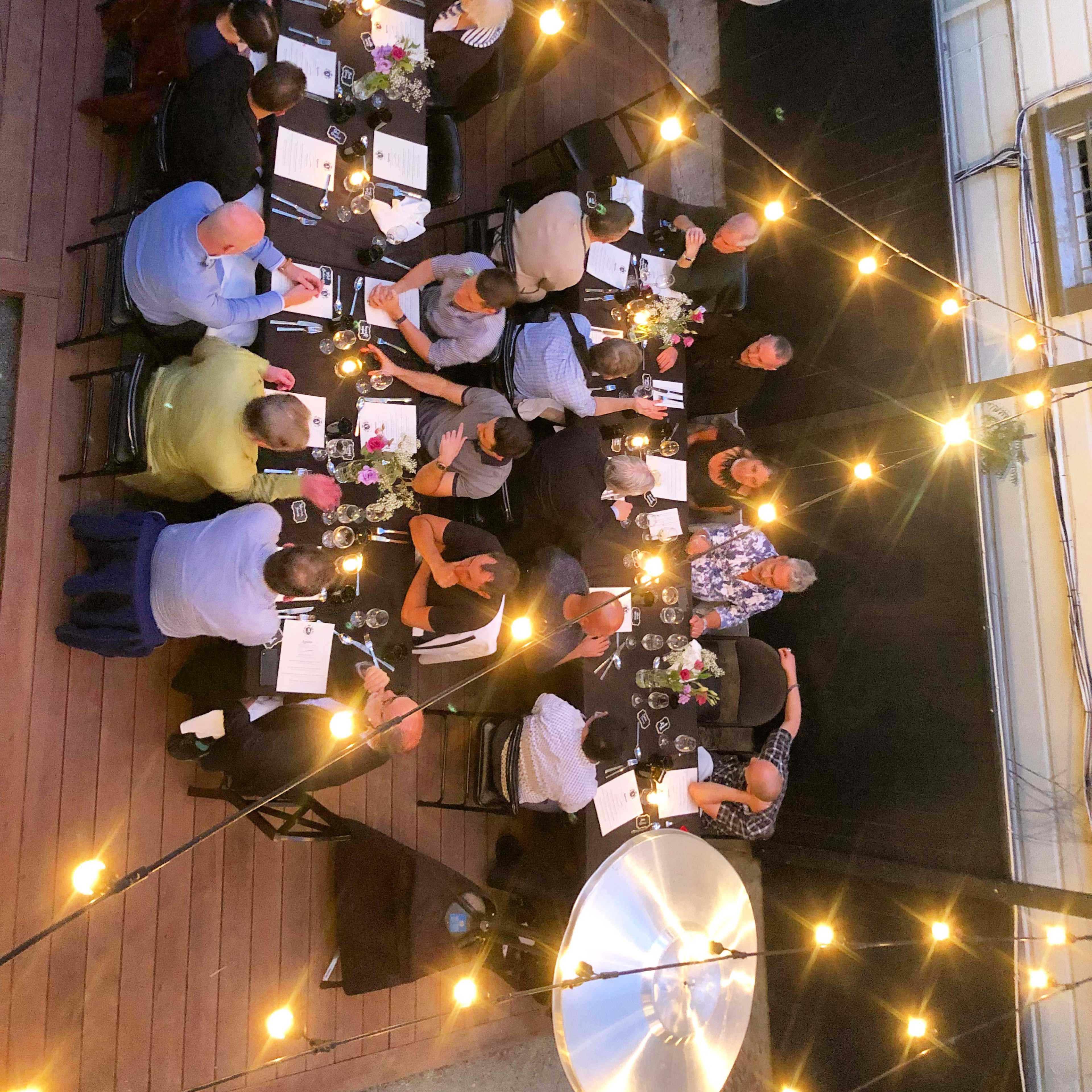 A long table is set for a dinner party outdoors, with guests seated and string lights illuminating the scene.