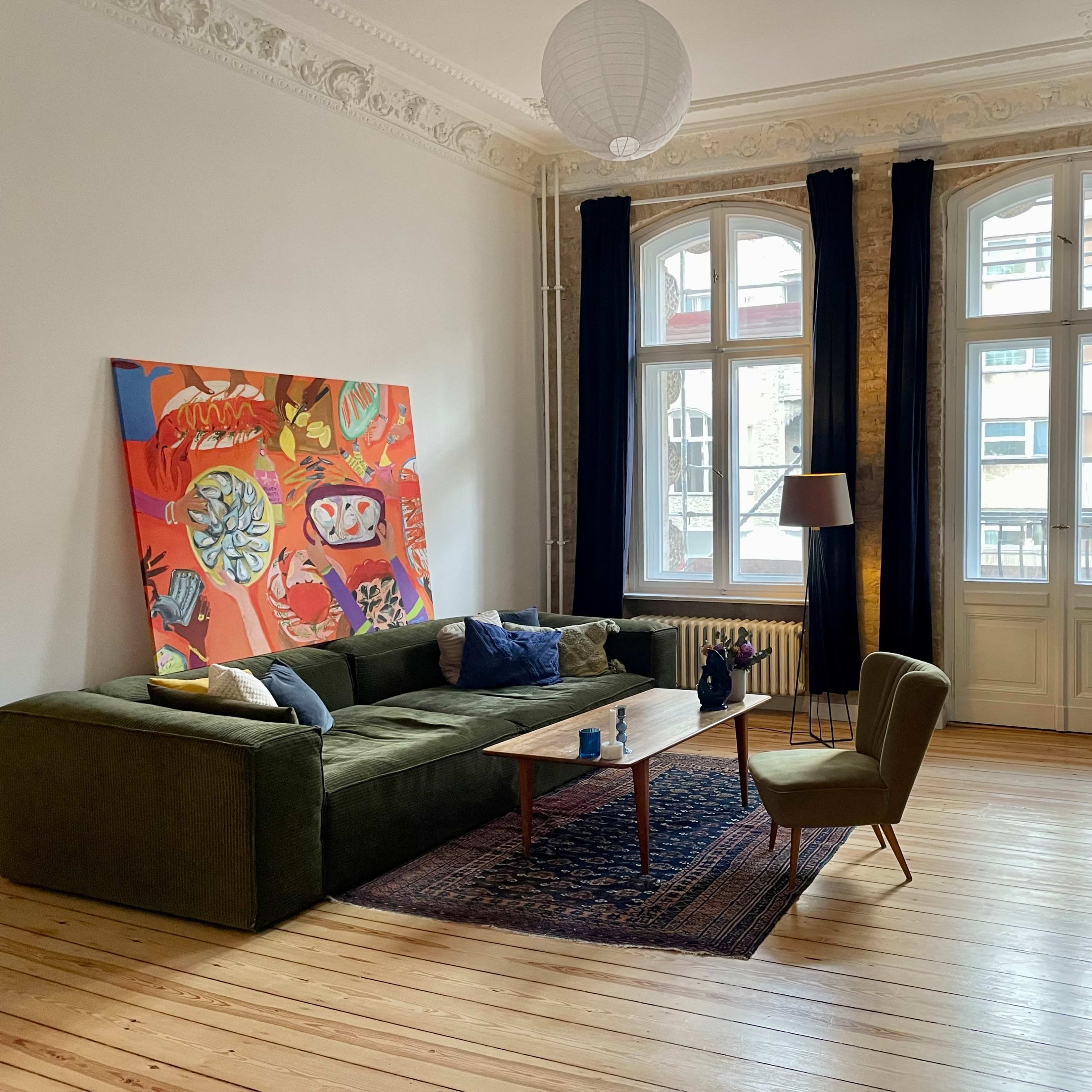 A bright living room with large windows, a green sectional sofa, a wooden coffee table, and an abstract painting on the wall.