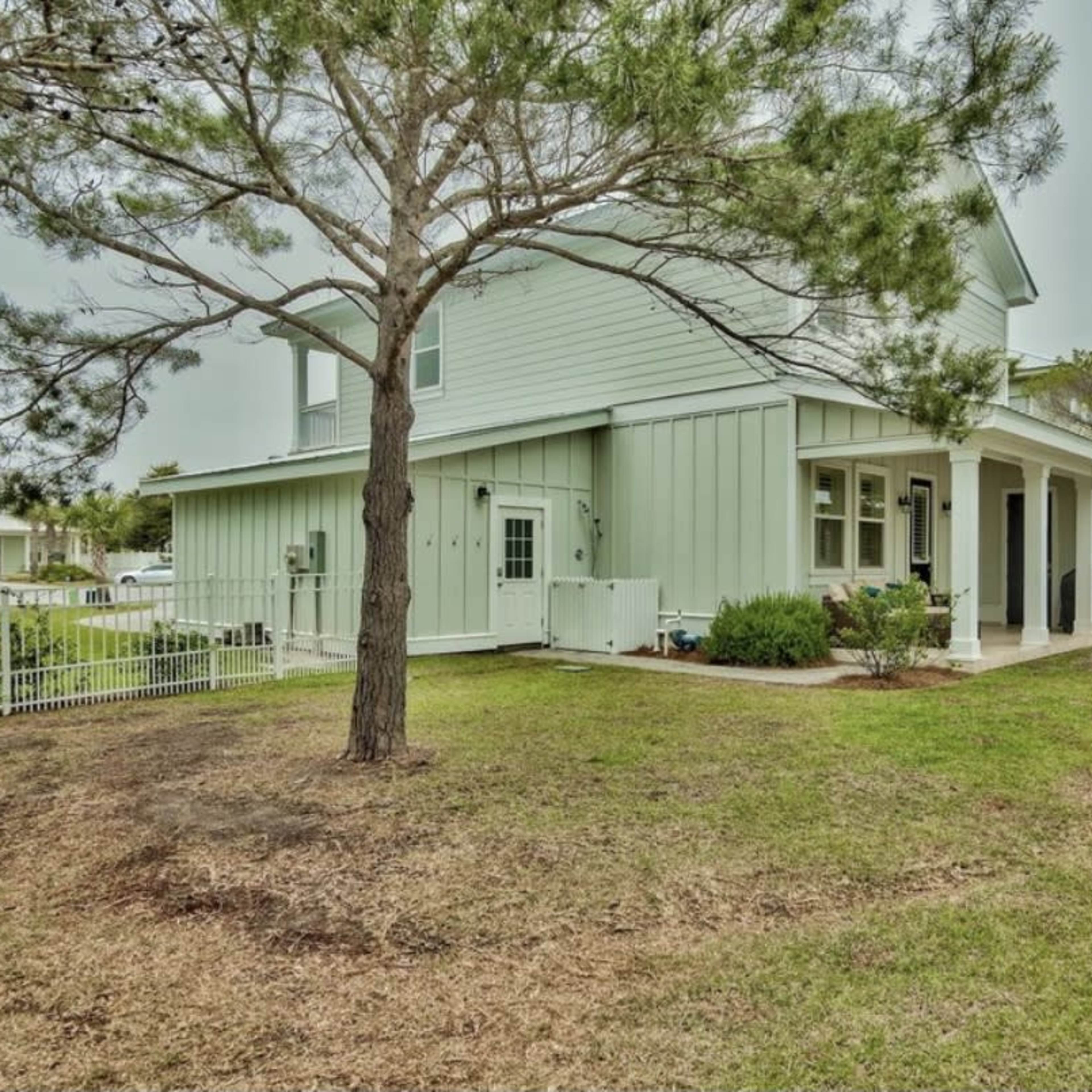 The side view of a two-story house surrounded by grass and a few trees, with a porch and a fenced yard.