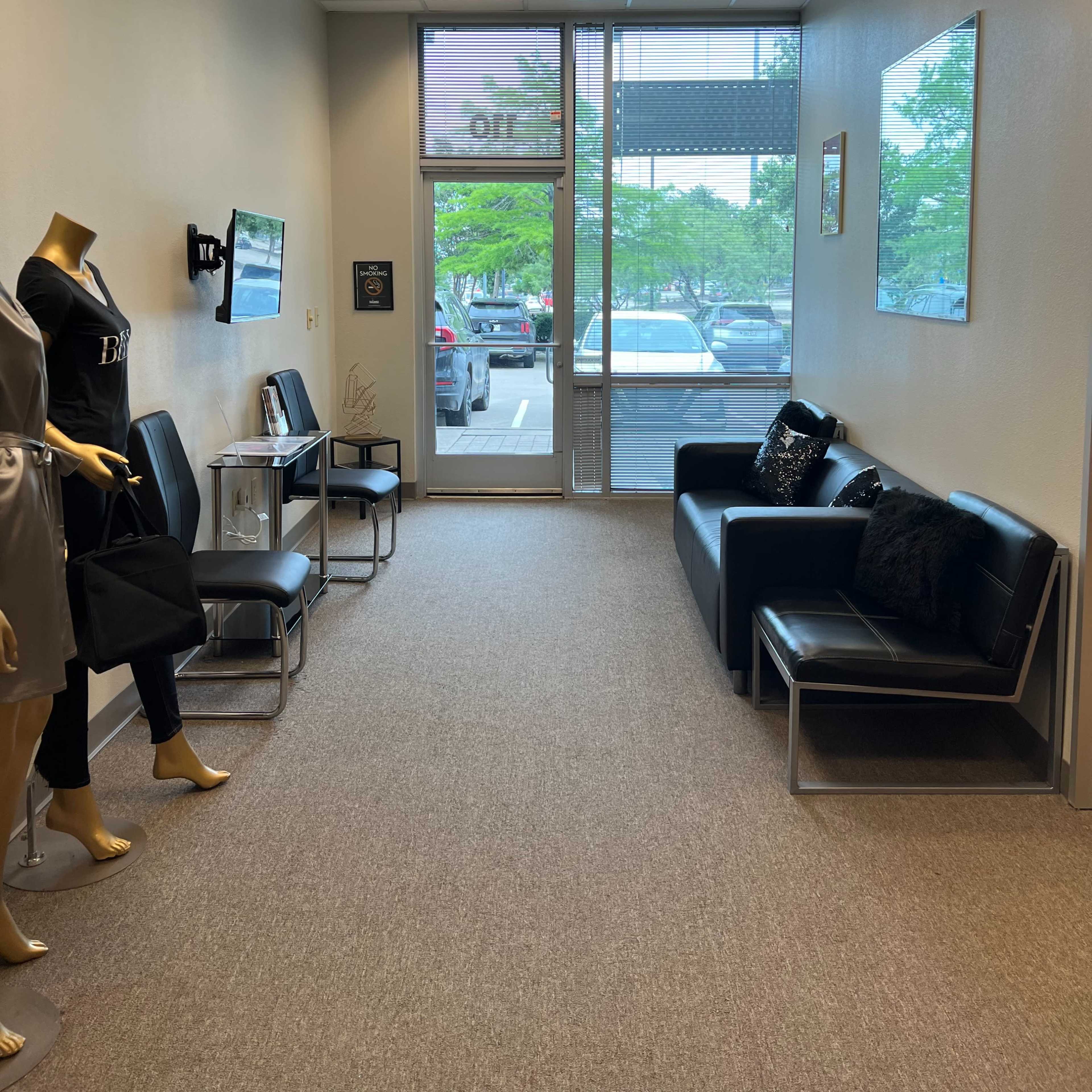 A narrow hallway with a seating area, a mannequin displaying clothing, and large windows that overlook a parking lot.
