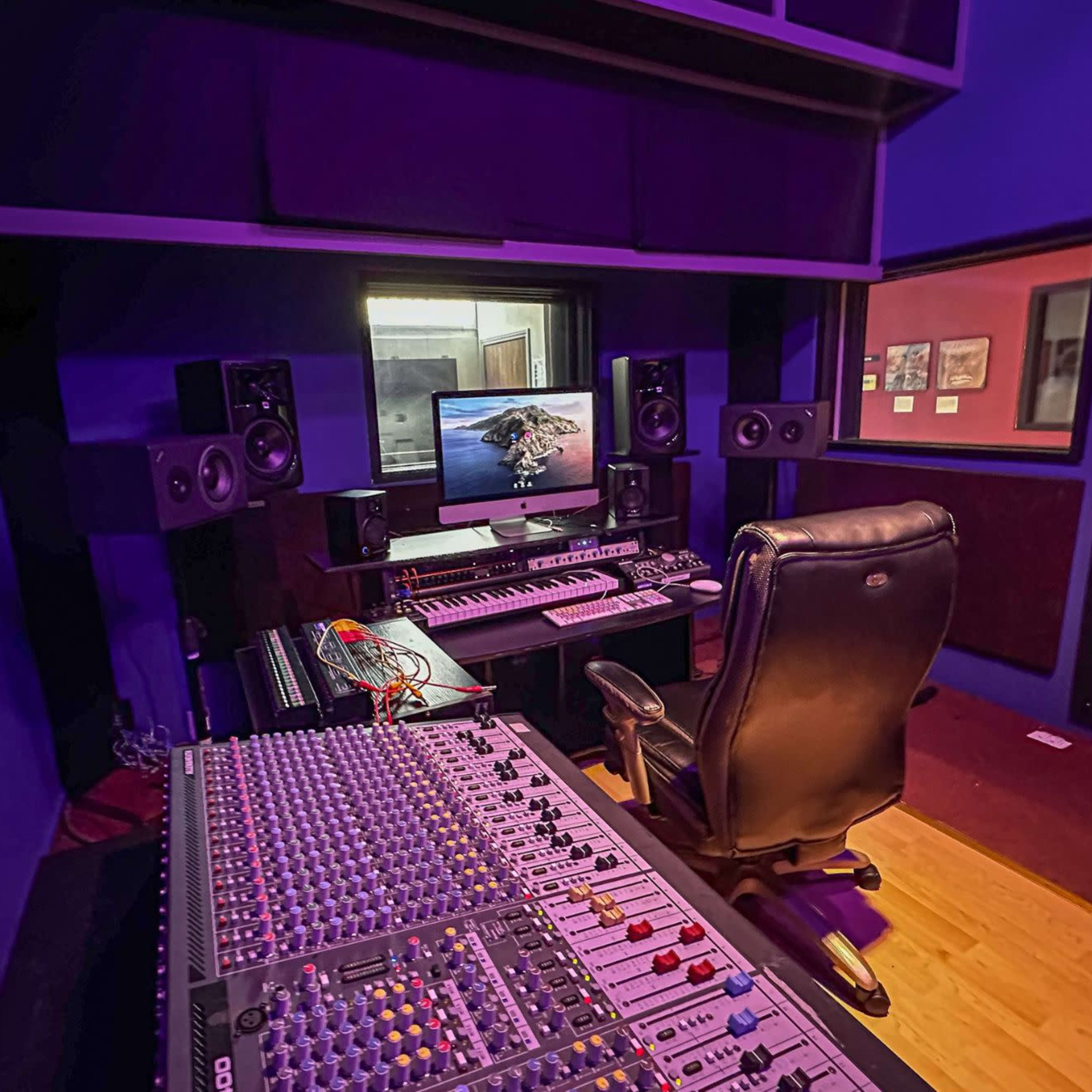 A music production studio featuring a mixing console, computer setup, speakers, and a comfortable chair, all within a purple-walled environment.