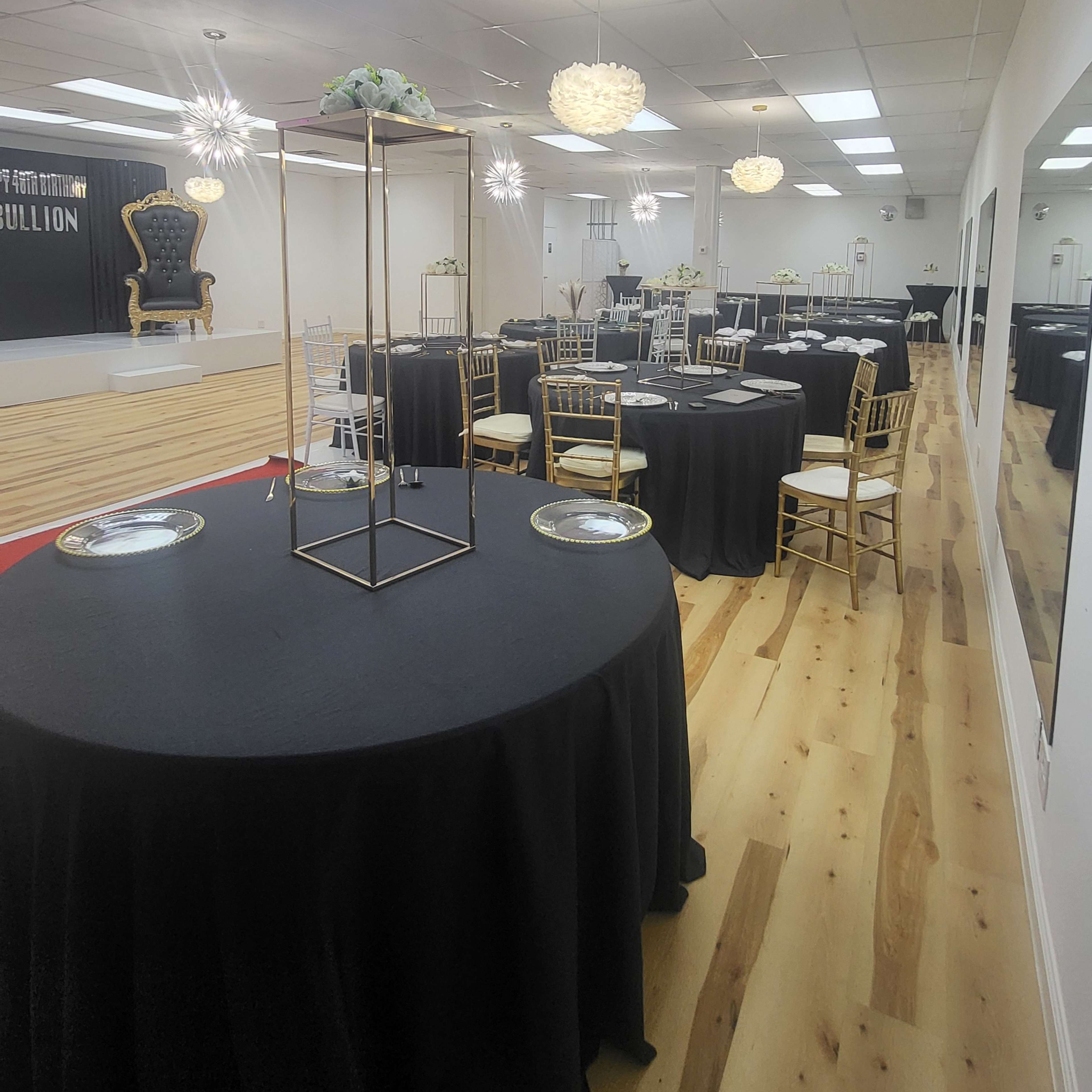 A spacious event hall with several round tables covered in black tablecloths, elegantly arranged with gold-framed centerpieces and decorative chairs.