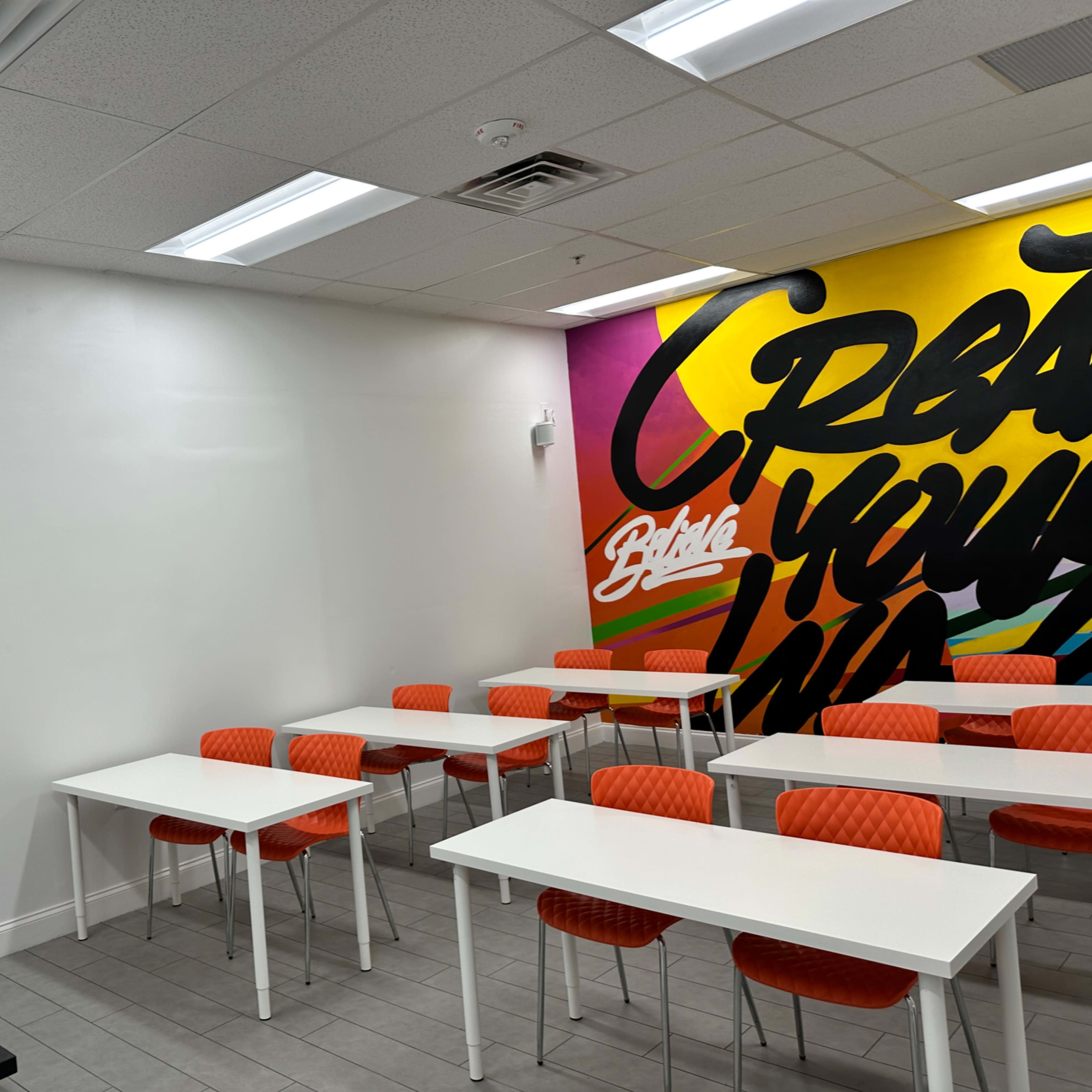 A bright classroom features orange chairs and white tables, with a colorful mural on one wall that reads "Create Your Own."