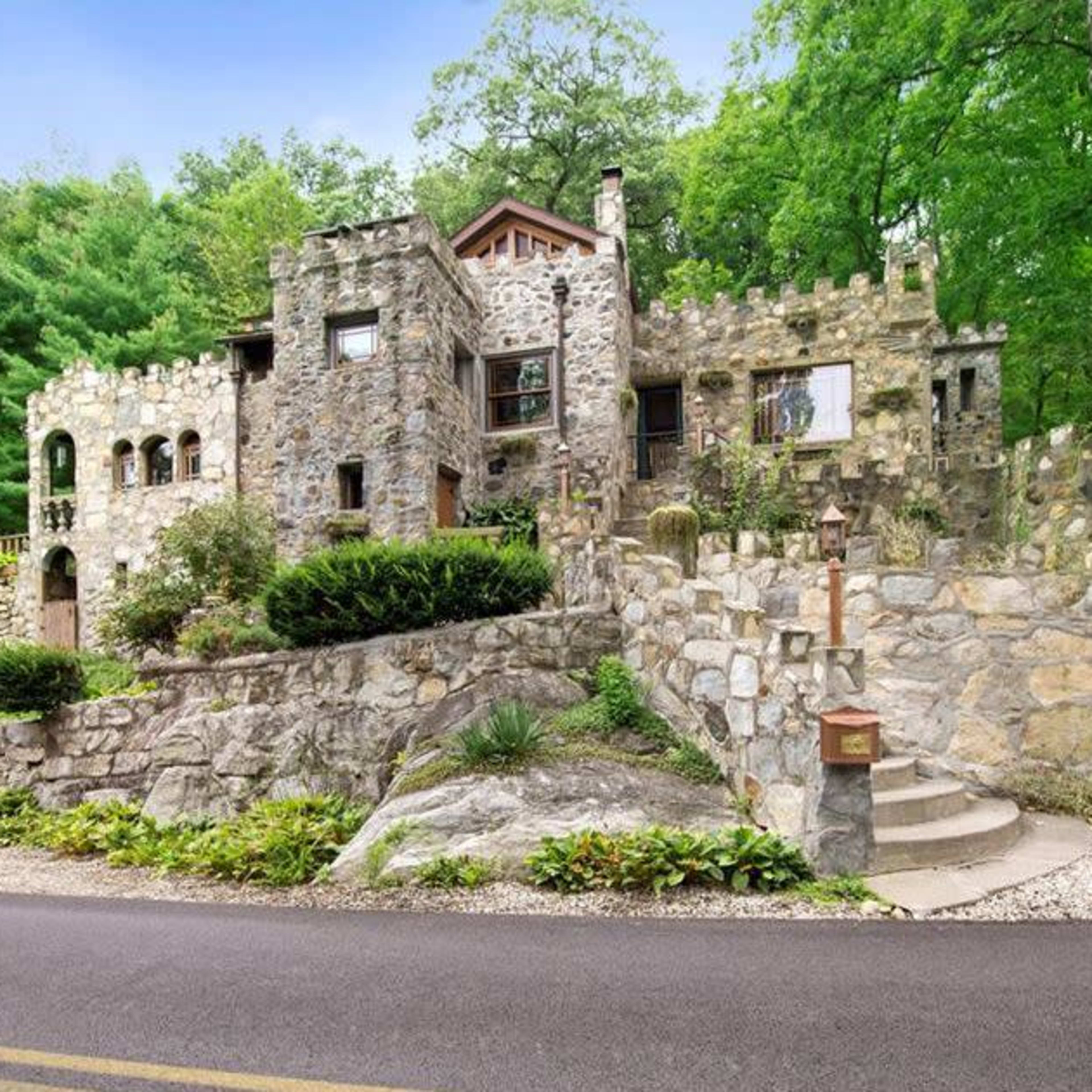 A stone castle-like house is set among trees, positioned on a hillside with a winding driveway leading up to it.
