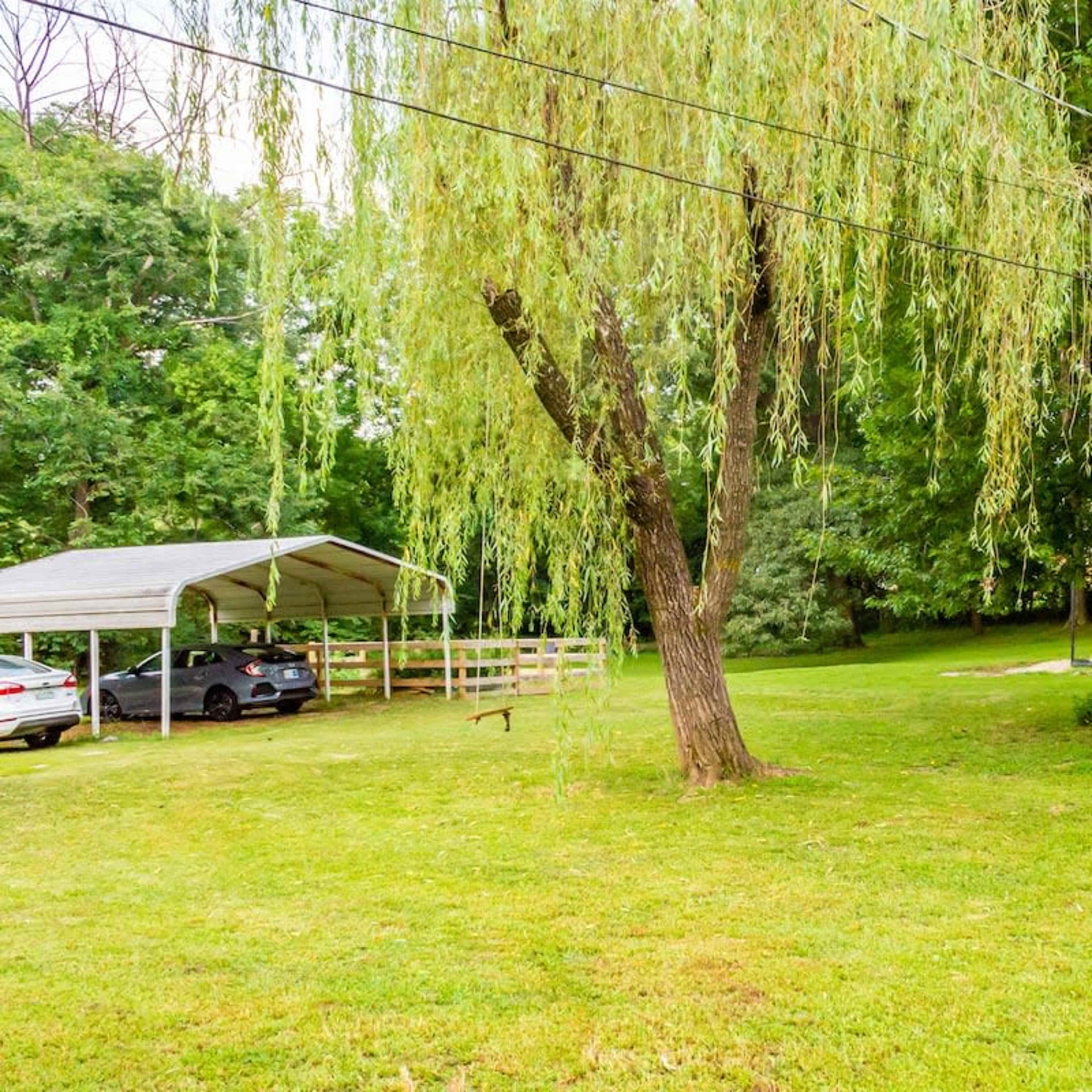 A small gray shed with a red door sits in a grassy area beside a covered carport with two vehicles underneath.
