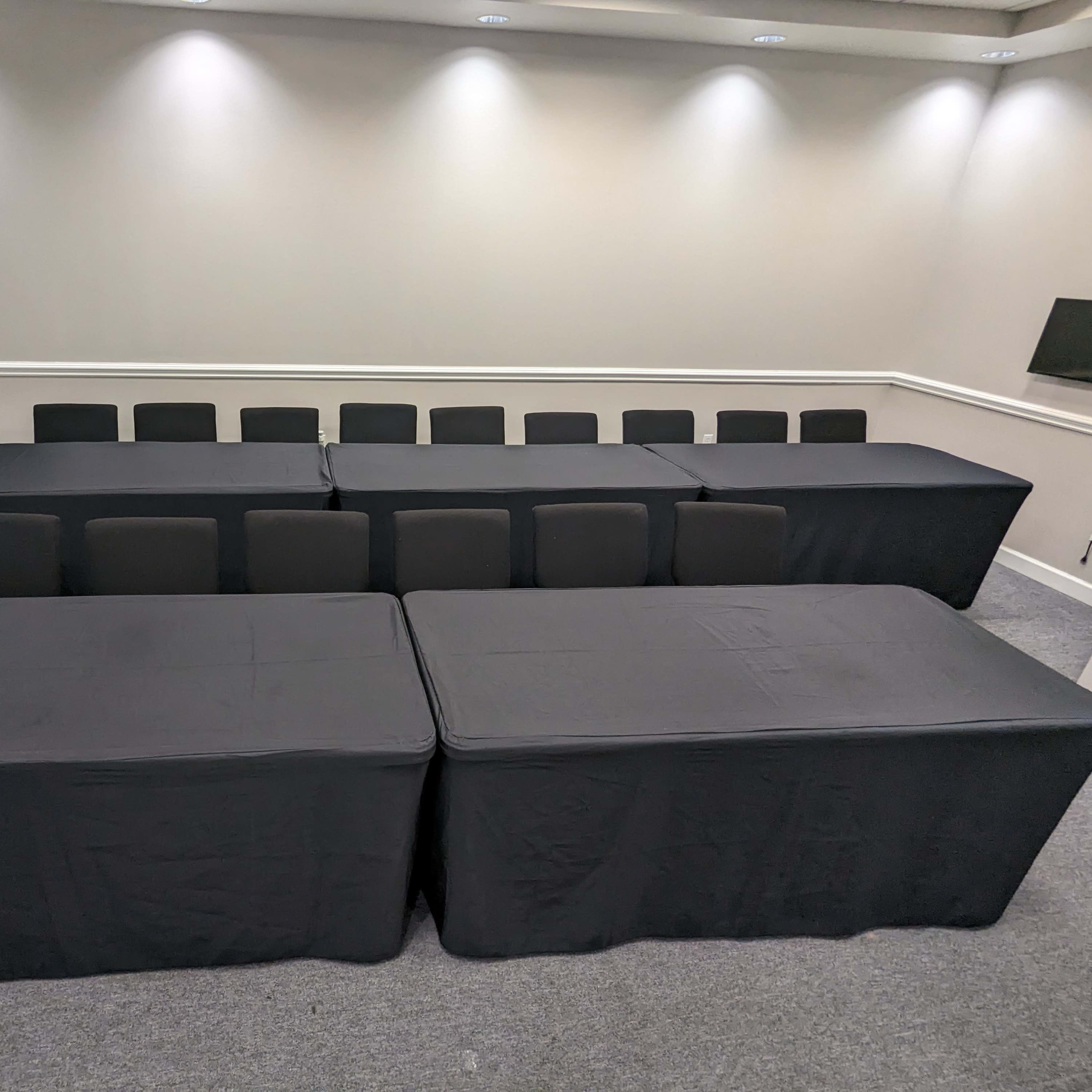 A conference room set up with multiple black-covered tables arranged in a rectangular formation, surrounded by black chairs.