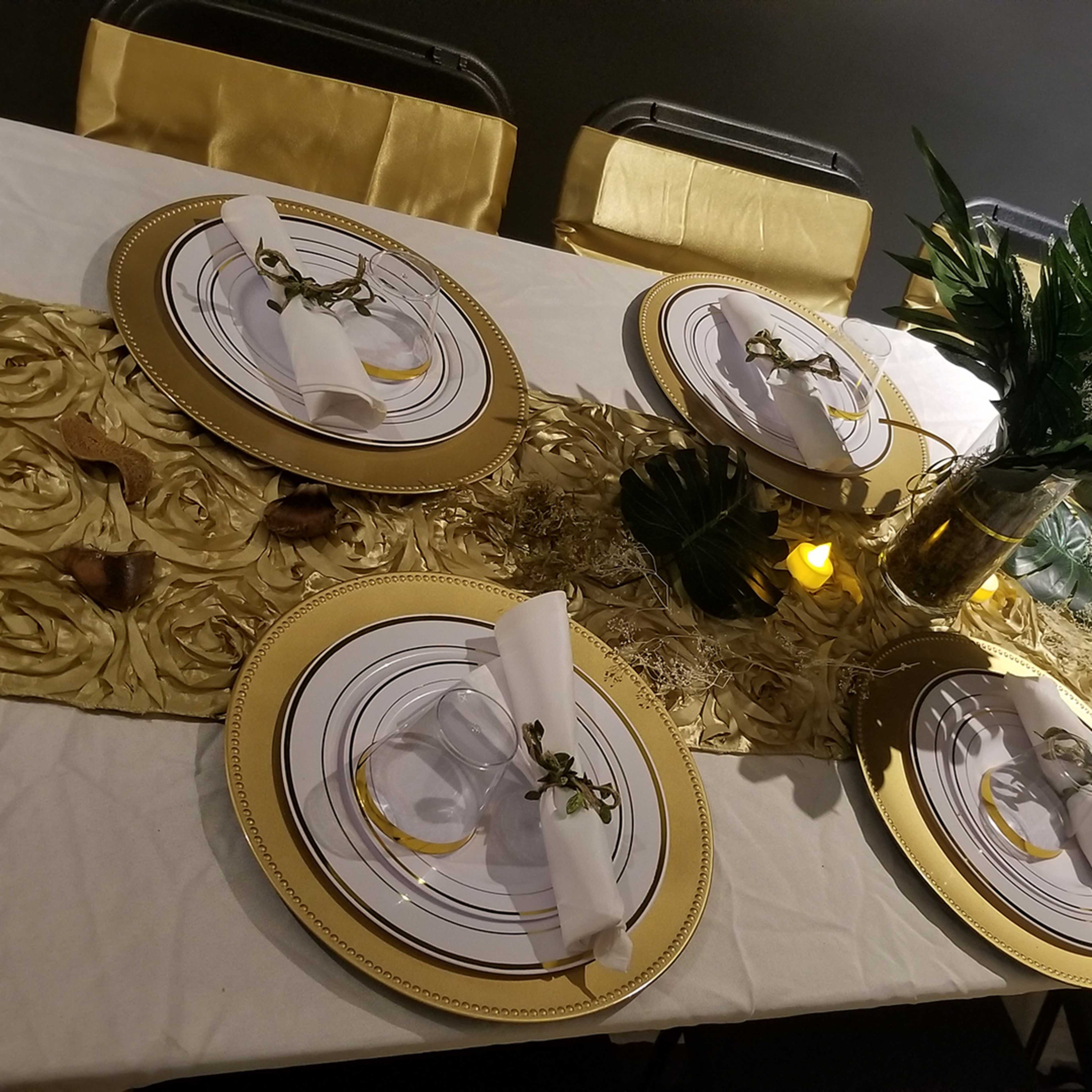 A dining table is set with gold-themed plates, napkins, and decorations, with a central arrangement of greenery and candlelight.