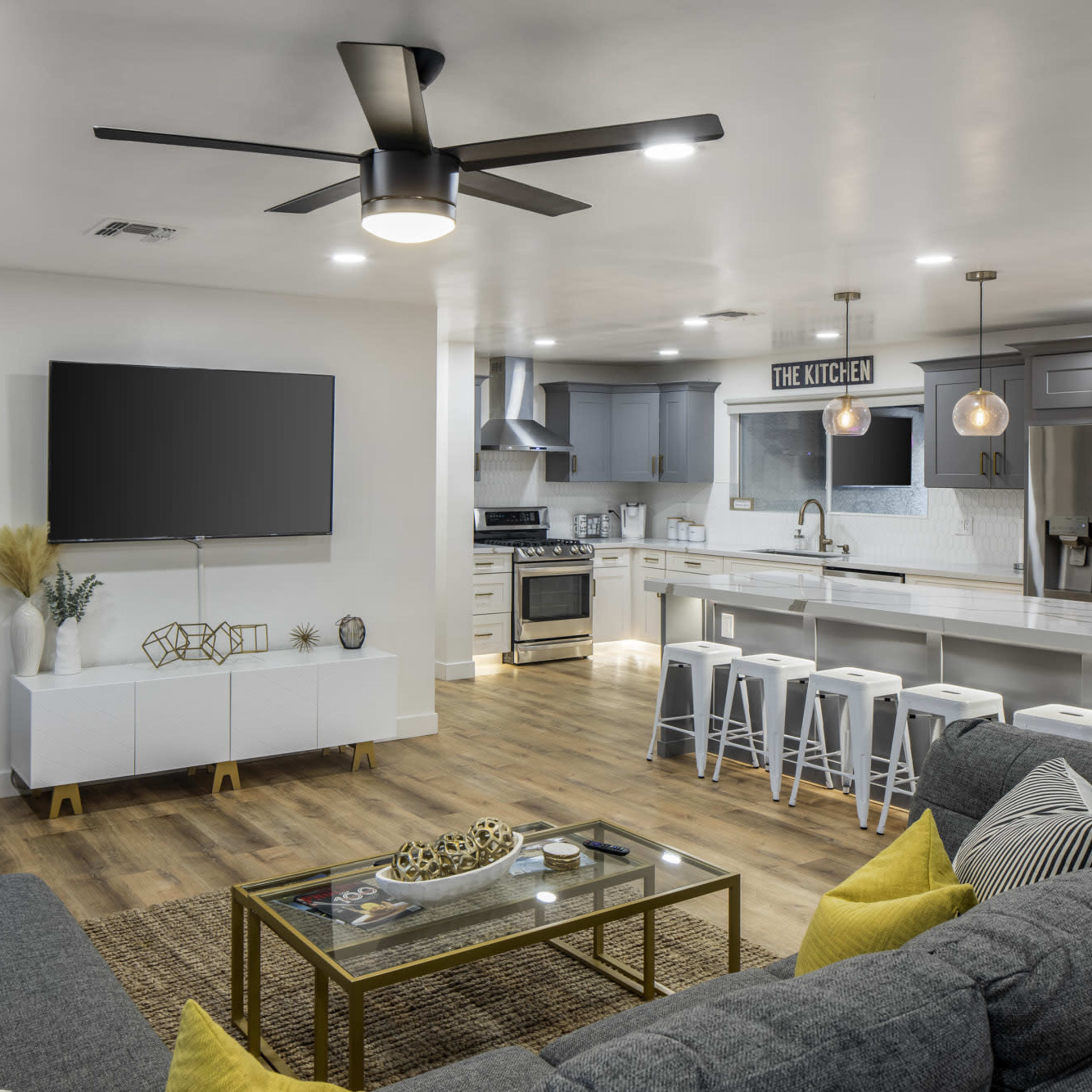 A modern living room and kitchen area with a ceiling fan, large TV, comfortable seating, and a sleek kitchen island with bar stools.