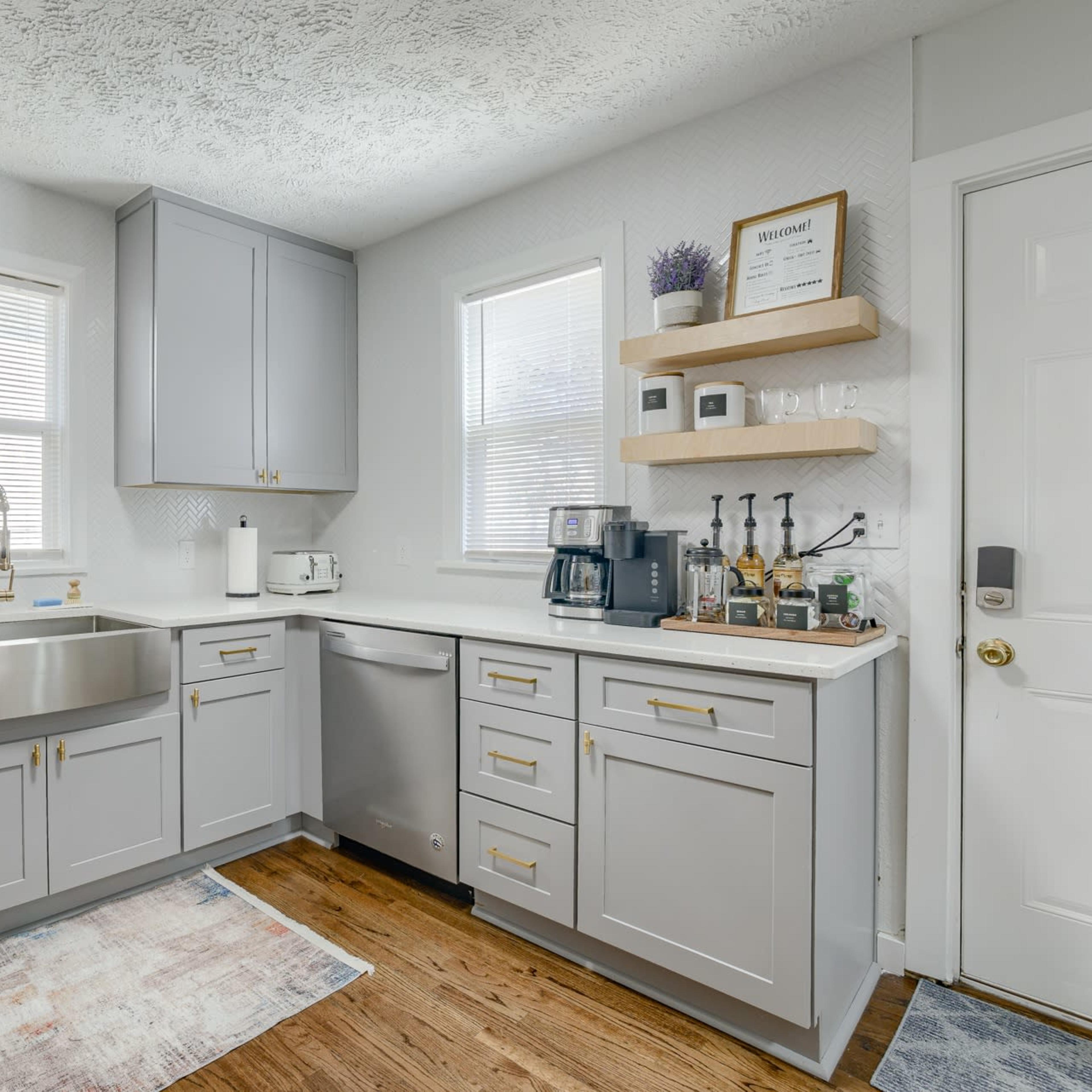 A modern kitchen features gray cabinets, a stainless steel sink, and a wooden floor, with a small shelf displaying kitchen items.