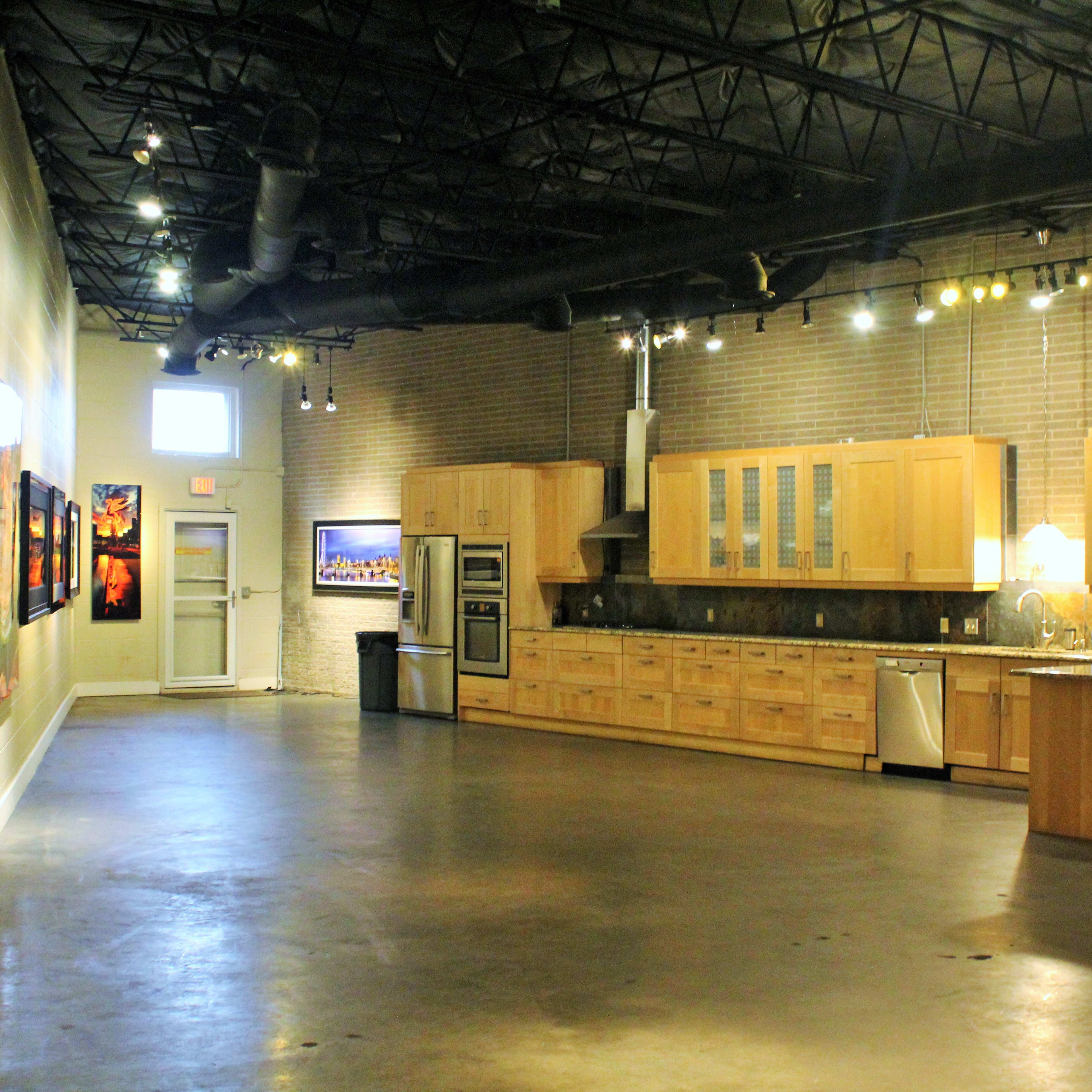 An open gallery space featuring a kitchen area with wooden cabinets and multiple framed artworks displayed on the walls.