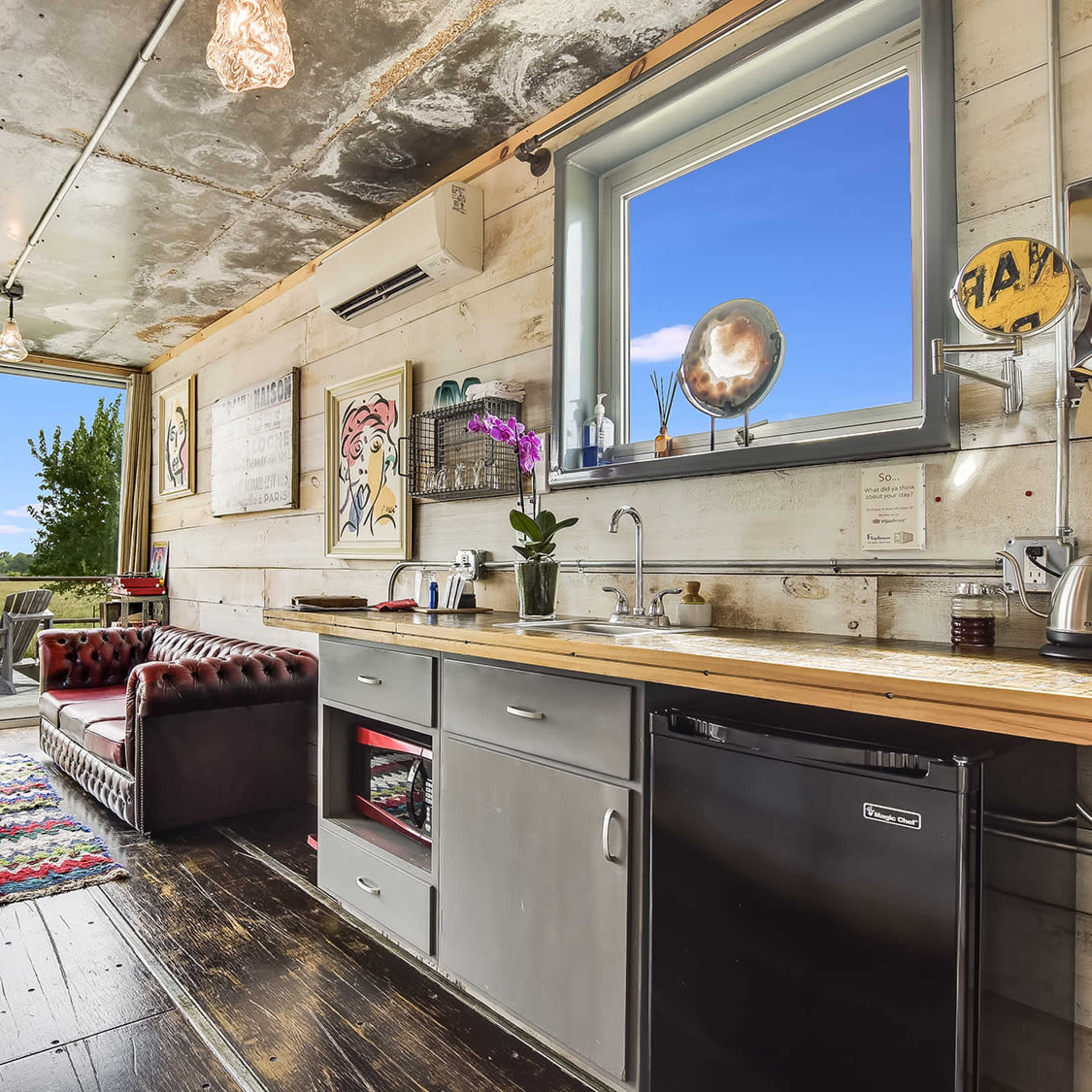 A small kitchen and living area with a sofa, wooden countertop, sink, and refrigerator, featuring a large window that overlooks a scenic landscape.