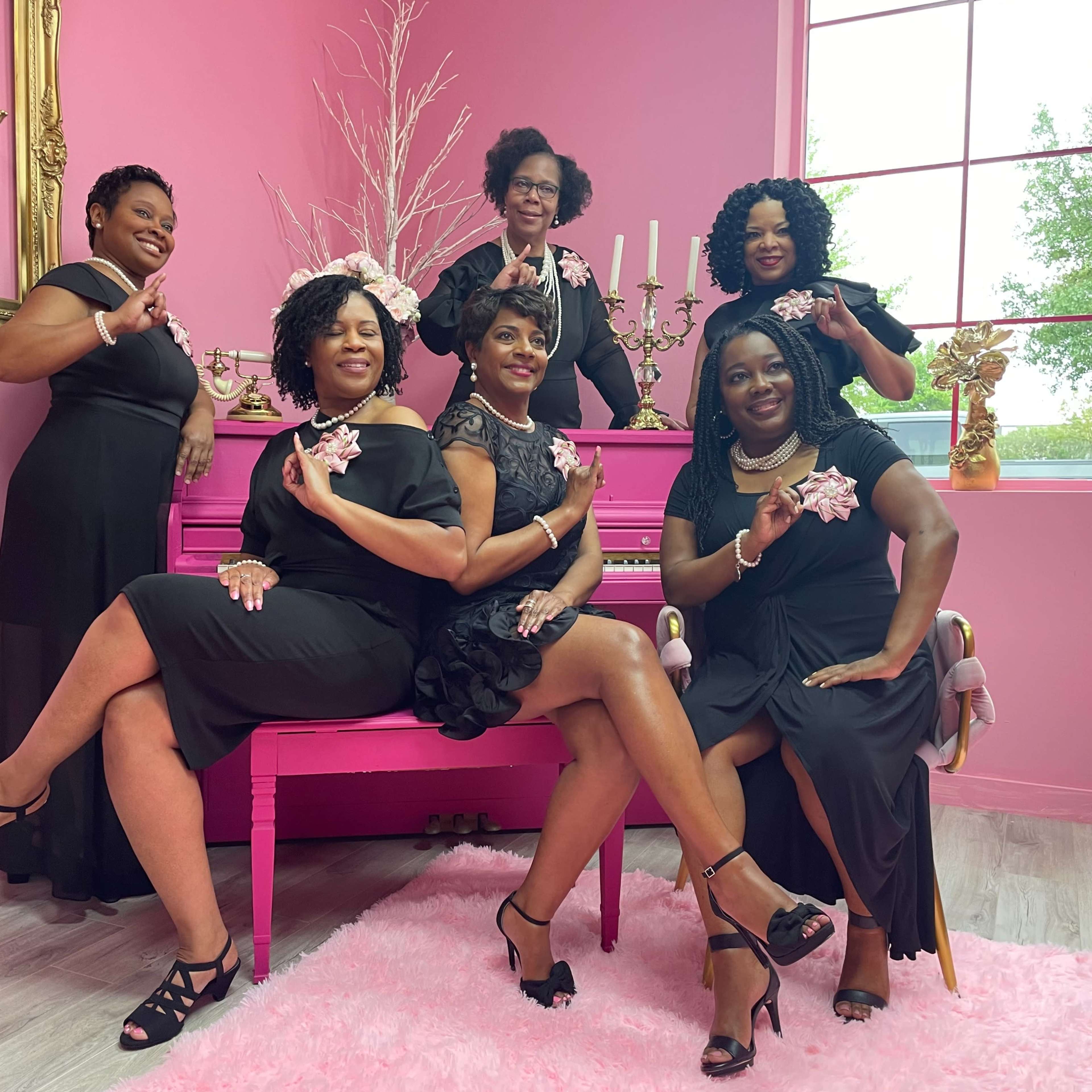 A group of six women in black dresses poses together in a pink themed room with a piano and decorative items.