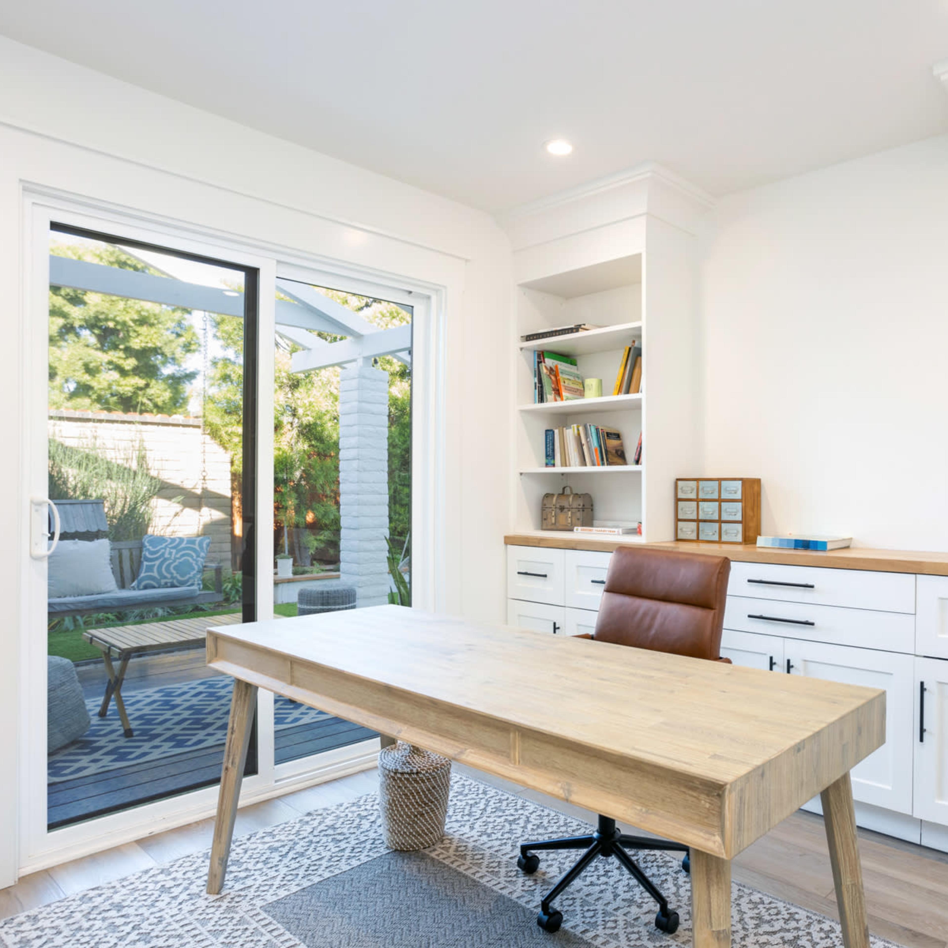 A modern home office features a wooden desk, a leather chair, and bookshelves filled with colorful books, while a glass sliding door provides a view of the outdoor area.