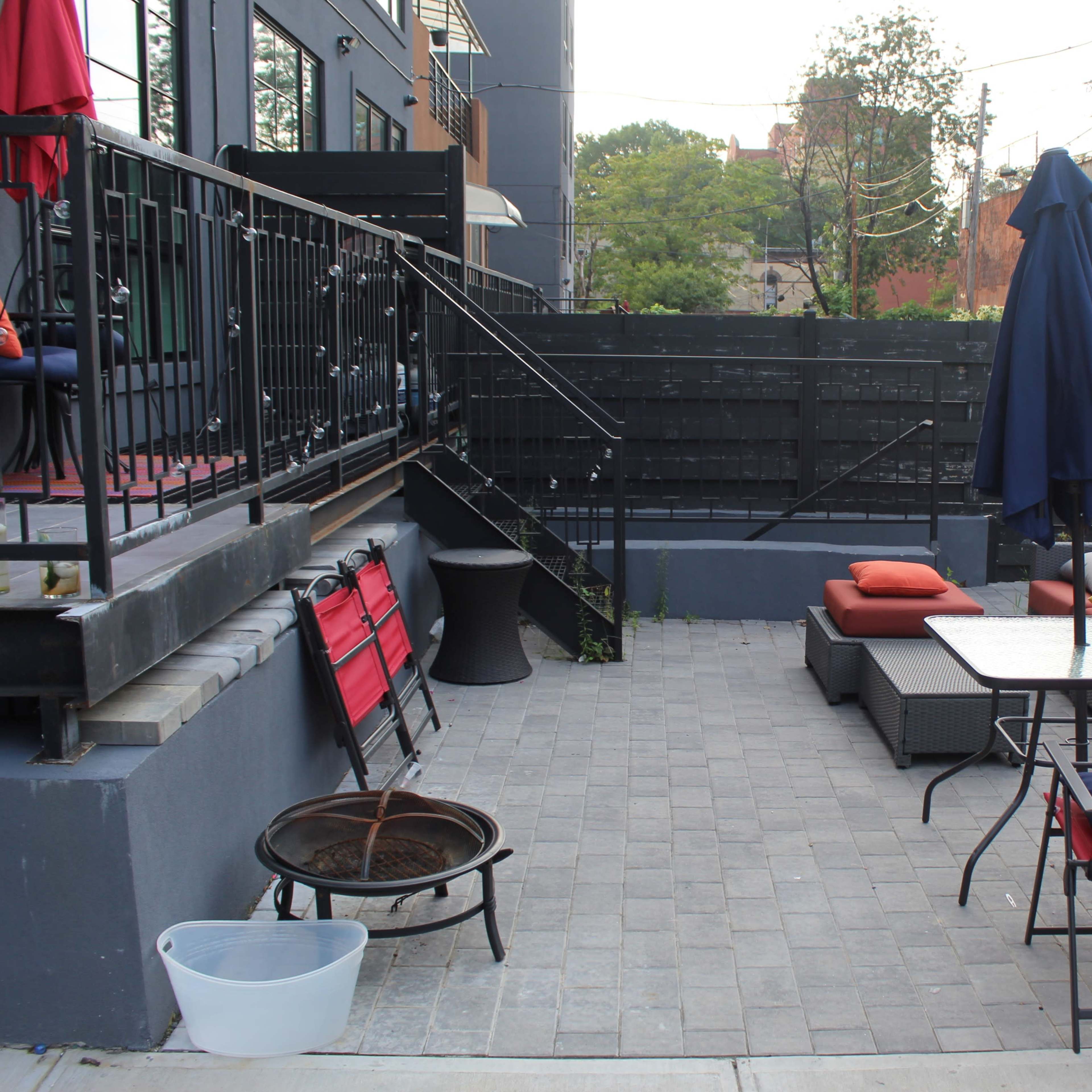 A patio area with a seating arrangement, an umbrella table, a fire pit, and red chairs alongside a black fenced wall.