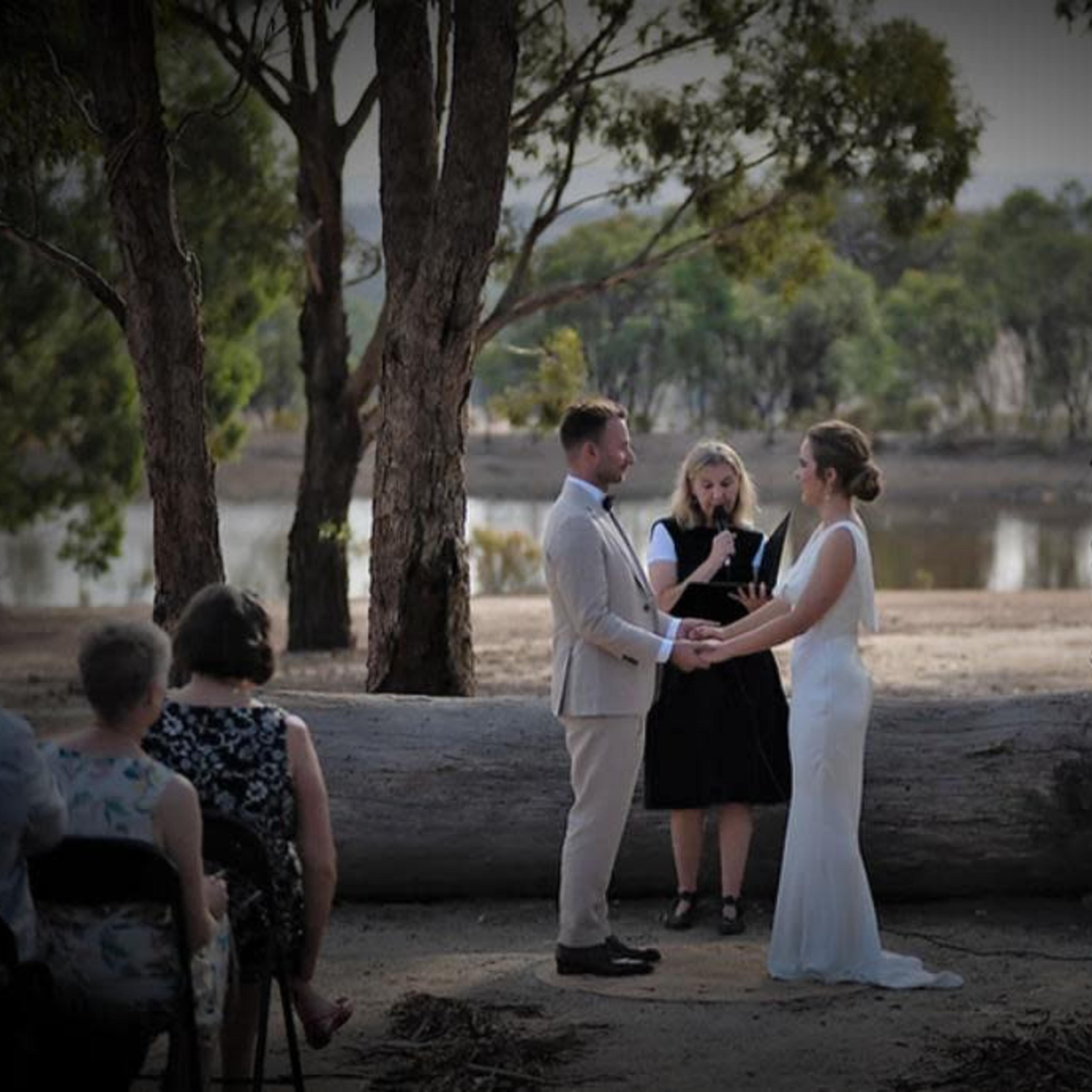 A couple exchanges vows during a wedding ceremony by a lake, with guests seated on folding chairs in the foreground.